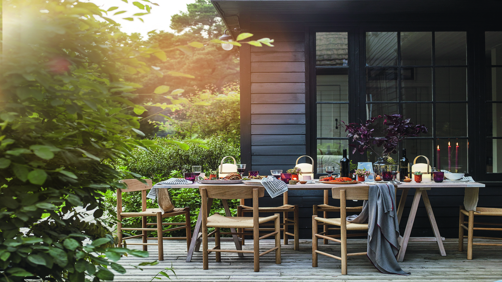 Eine holzgetäfelte Terrasse vor einem holzgetäfelten Haus mit einem großen Esstisch mit Tischdekoration, Holzstühlen und einem großen Busch im linken Vordergrund.