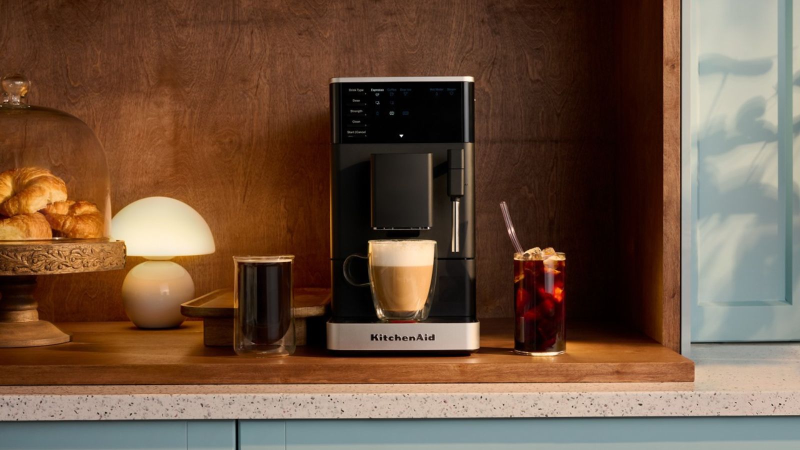 Eine schwarze, kompakte KitchenAid-Kaffeemaschine auf einer Holztheke mit Holzrückwand, daneben eine Lampe und ein Kuchenständer voller Gebäck. 