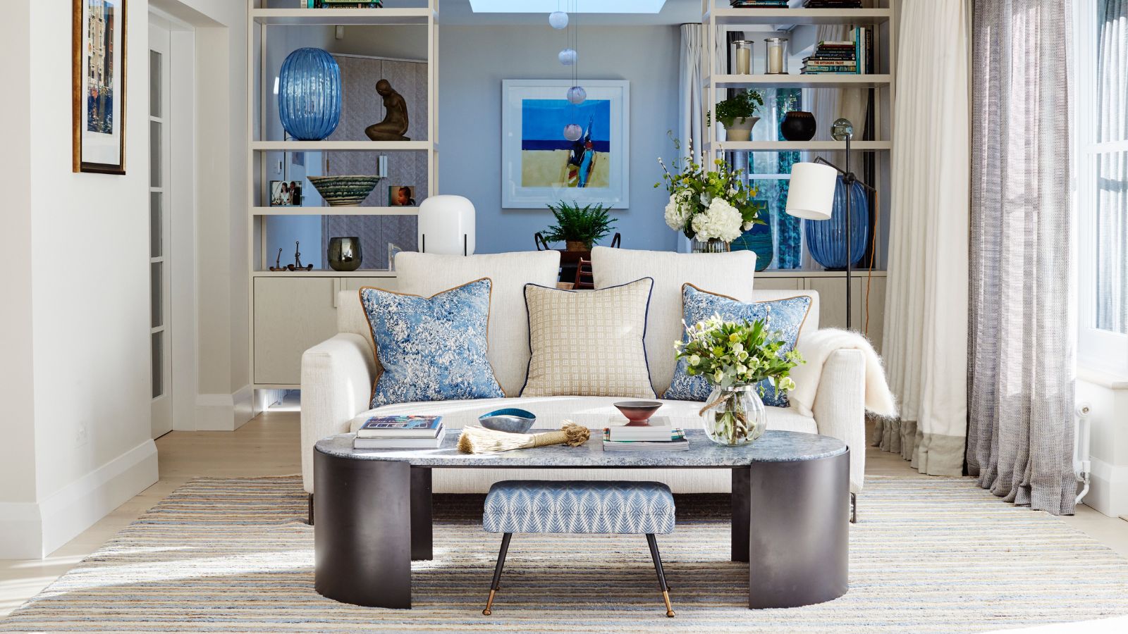 Ein helles Wohnzimmer mit einem ovalen schwarz-grauen Couchtisch und einem kleinen weißen Sofa mit blauen und weißen Kissen in der Mitte, mit weißen Wänden mit weißen Vitrinen und einem hellgrauen Teppich. Auf der rechten Seite befindet sich ein großes Fenster mit mehrschichtigen braunen, durchsichtigen Vorhängen und weißen Vorhängen.