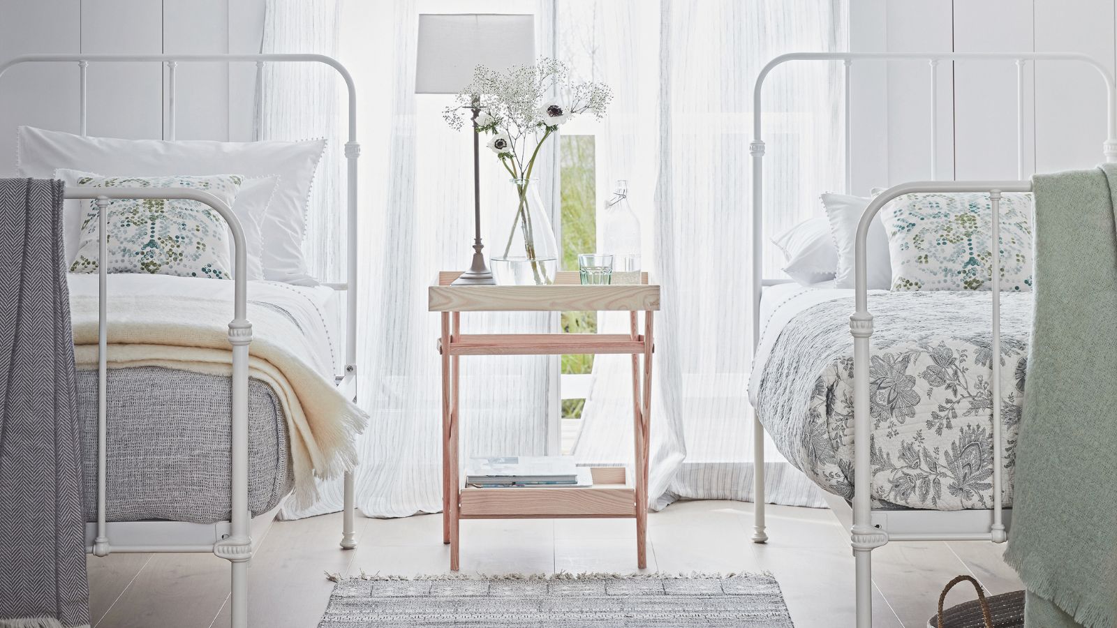 Ein strahlend weißes Schlafzimmer mit weißen Betten, einem großen Fenster mit transparenten Vorhängen, hellem Holzboden, einem grauen Teppich, einer weißen Lampe und einer mit Blumen gefüllten Glasvase.