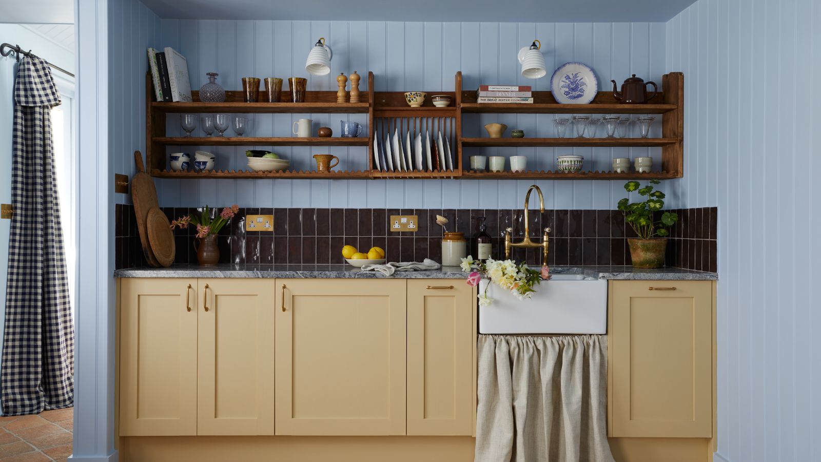 Eine gemütliche Landhausküche mit puderblau getäfelten Wänden und buttergelben Küchenschränken. Offene Holzregale und dunkle burgunderrote Rückwandfliesen. 