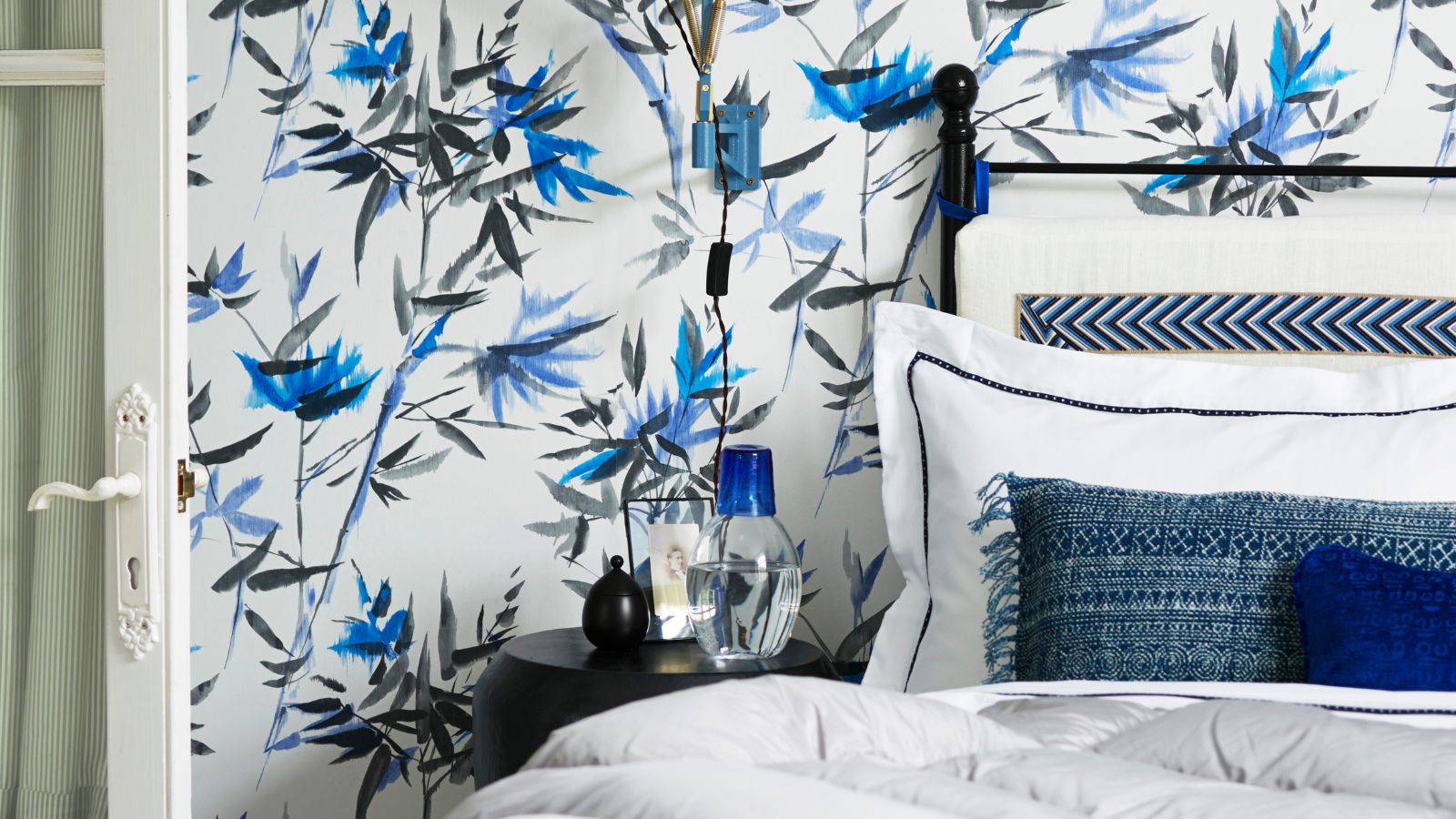 Blaues, weißes und schwarzes Schlafzimmer mit gemusterter Tapete, schwarzem Bett, weißer Bettwäsche, blauen Kissen, schwarzem Nachttisch und einer weißen offenen Tür auf der linken Seite.
