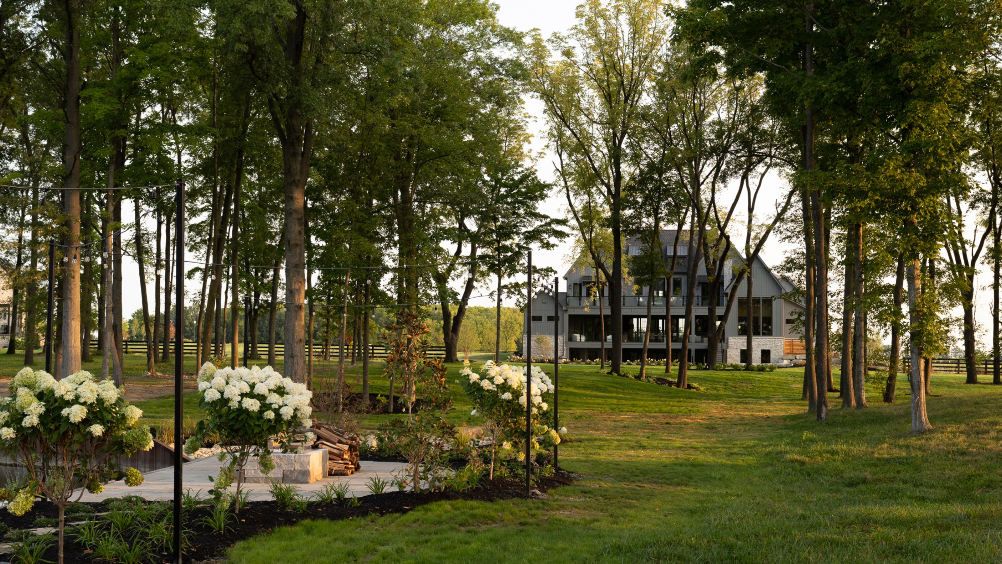 Eine Totalaufnahme eines modernen, mehrstöckigen Hauses im Bauernhausstil, eingebettet hinter einem Hain aus hohen Bäumen, mit einem gepflegten grünen Rasen und weiß blühenden Hortensien im Vordergrund.