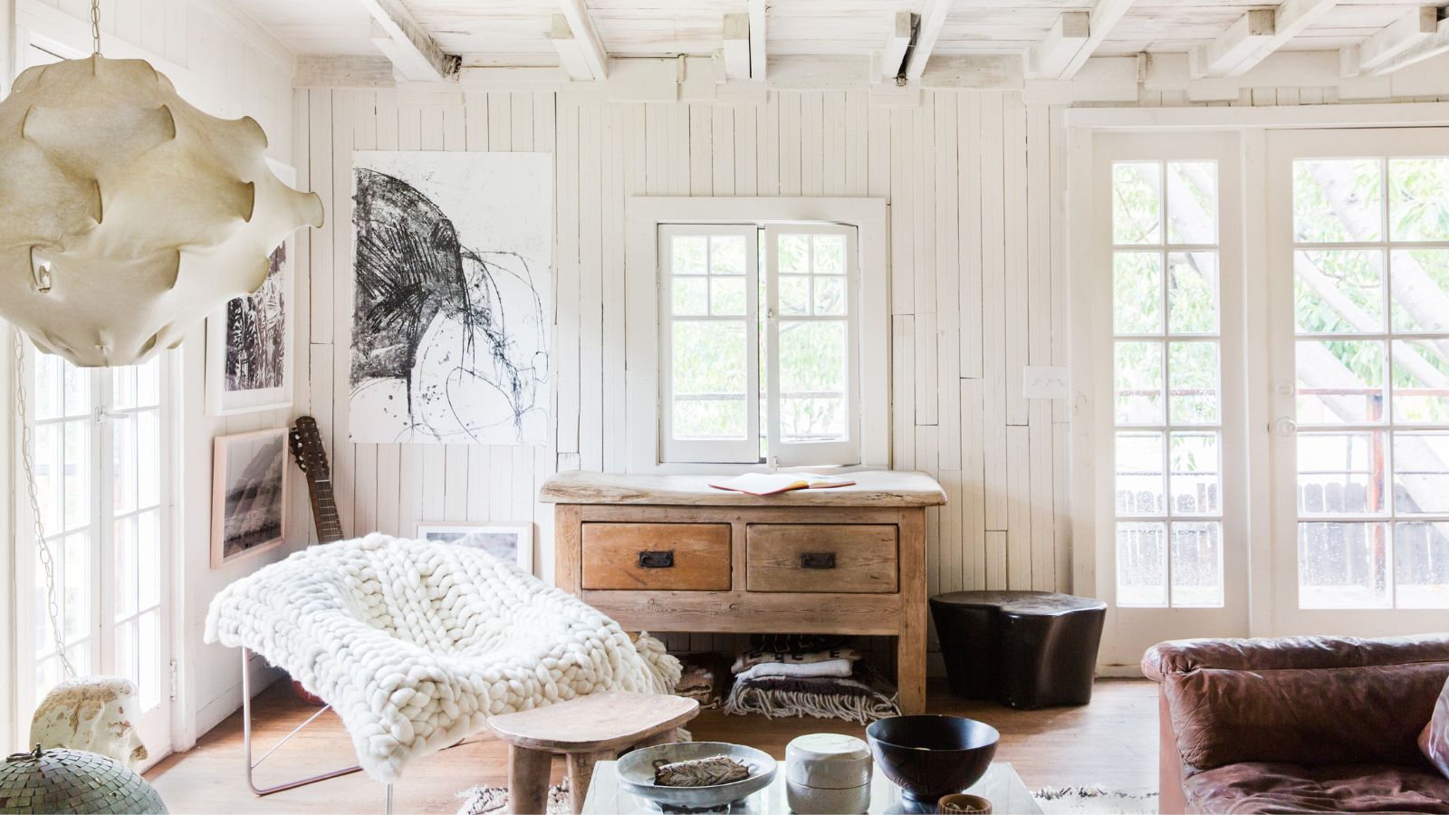 Wohnzimmer mit weißen Stufenwänden, französischen Türen, weißer Balkendecke, Holzboden, einer Holzkonsole unter dem Fenster, einem Metallsessel mit einem grob gestrickten Überwurf darüber und einem Ledersofa