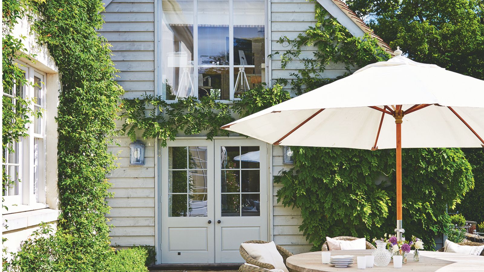 Eine sonnige Terrasse mit einem runden Tisch mit weißem Sonnenschirm, vor einem weiß getäfelten Haus mit großen Fenstern und Türen und überall grünen Kletterpflanzen.