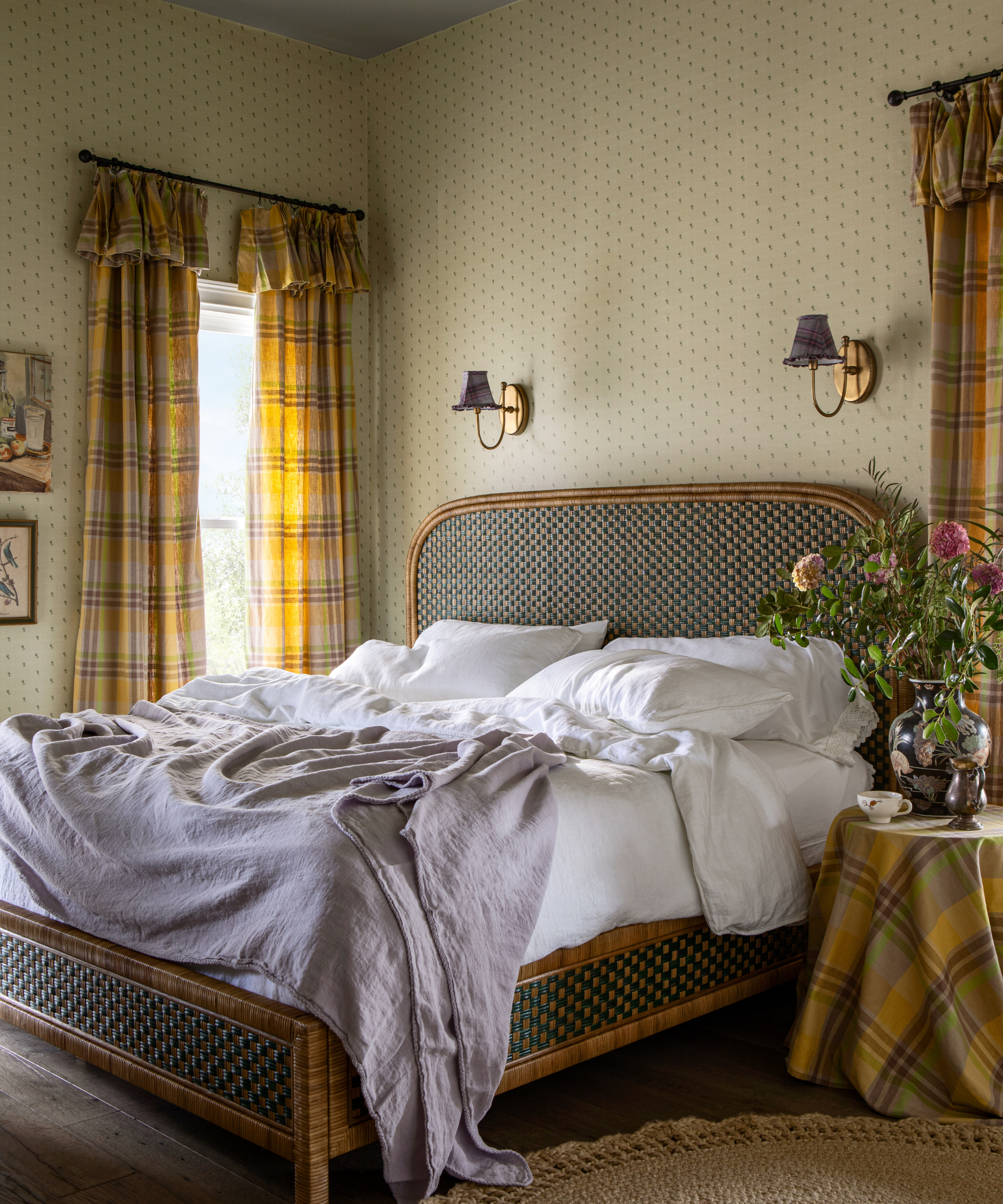 ein gemütliches Schlafzimmer mit winzigen Blumenmotiven auf der Tapete, einem gewebten Bettgestell und karierten bedruckten Vorhängen in Gelb und Braun
