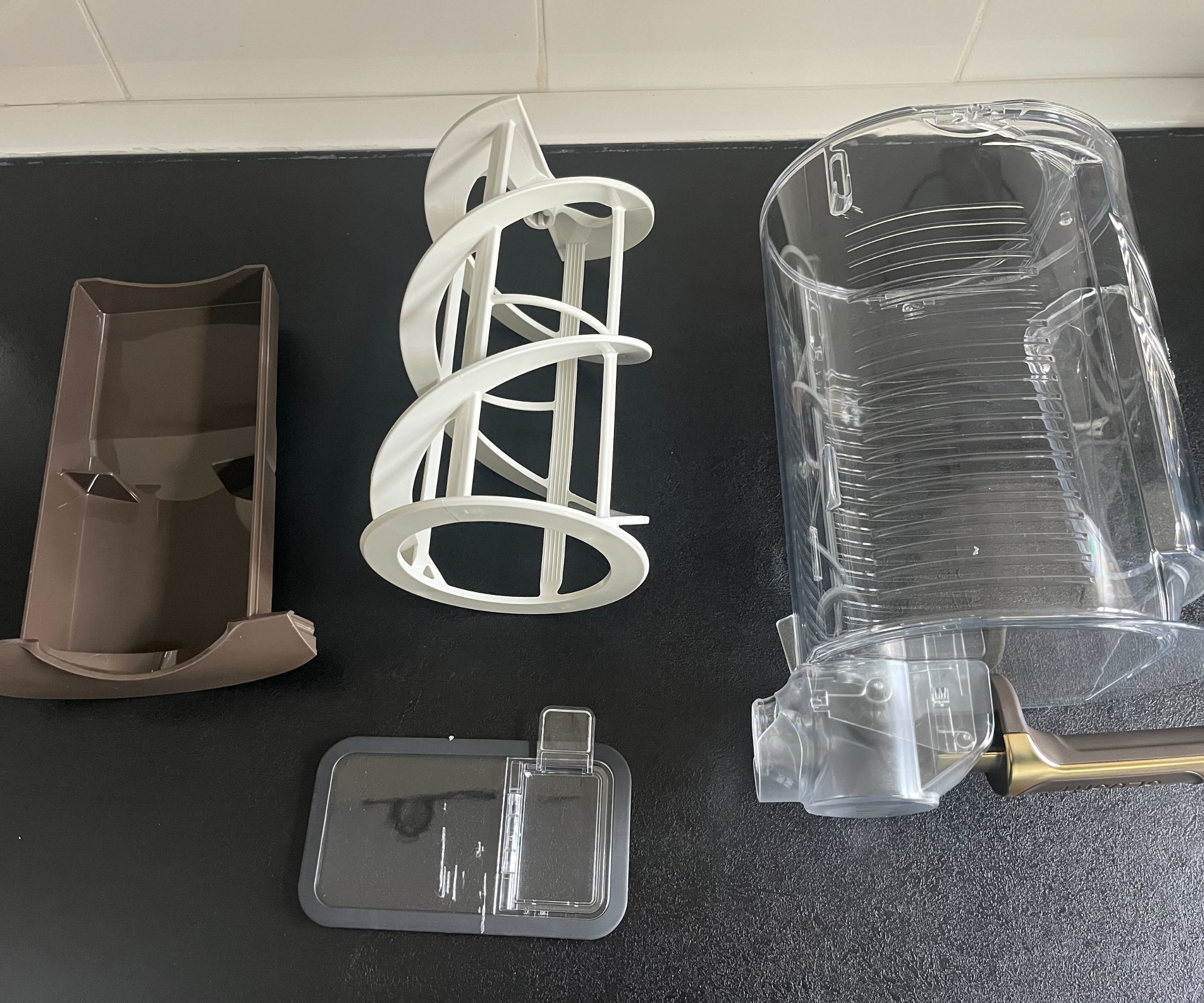 Die einzelnen Komponenten der Mischeinheit eines Ninja Slushi Max, mit dem durchsichtigen Behälter, der braunen Kondensationsschale, den Shite Paddles und dem Plastikdeckel, auf einer schwarzen Küchentheke. 