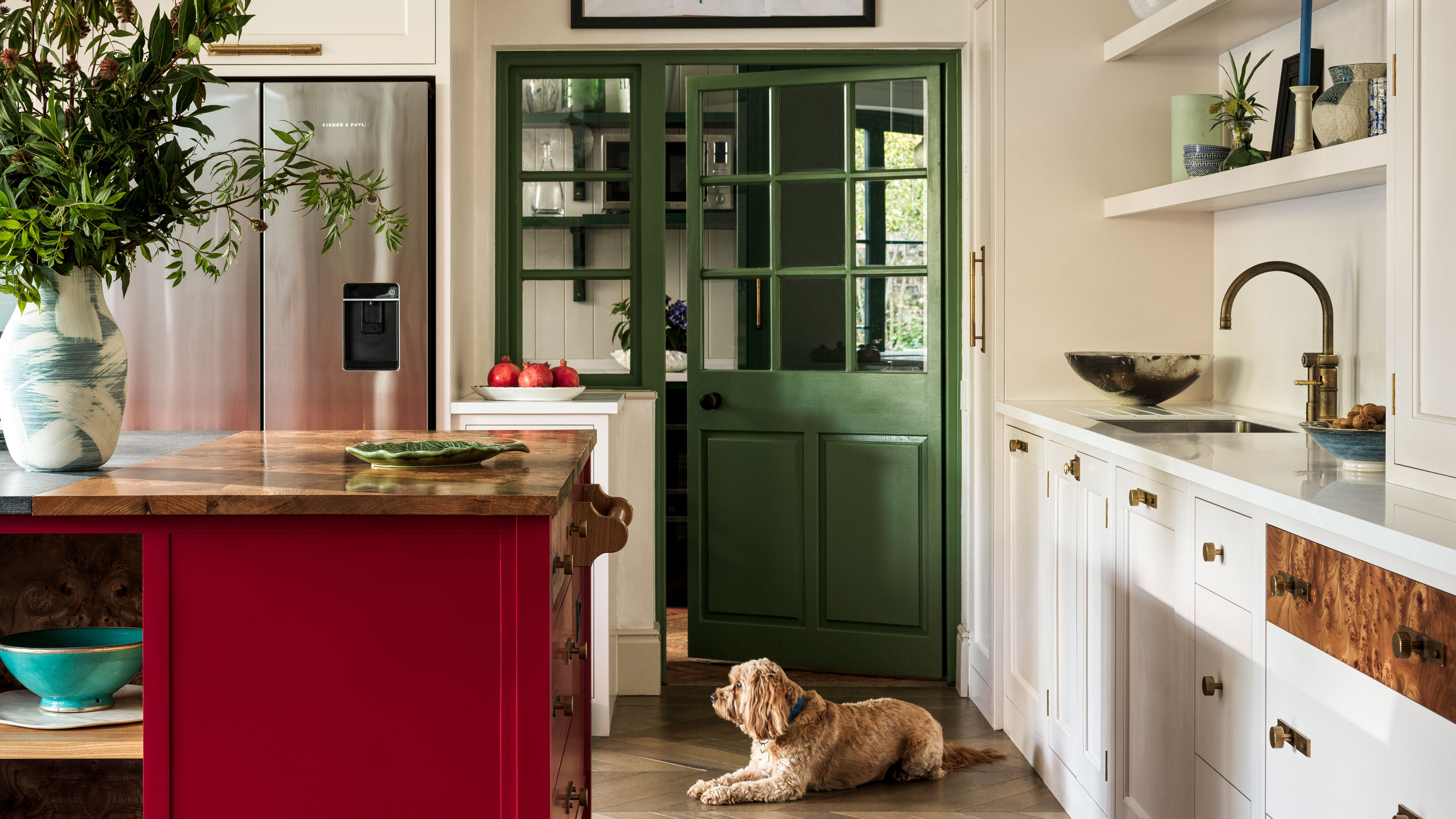 Eine helle Küche mit weißen Schränken, einer kräftig roten Kücheninsel mit Holzplatte und einer grün verglasten Tür, in der ein kleiner, flauschiger brauner Hund auf dem Boden liegt und auf einen Edelstahlkühlschrank blickt.