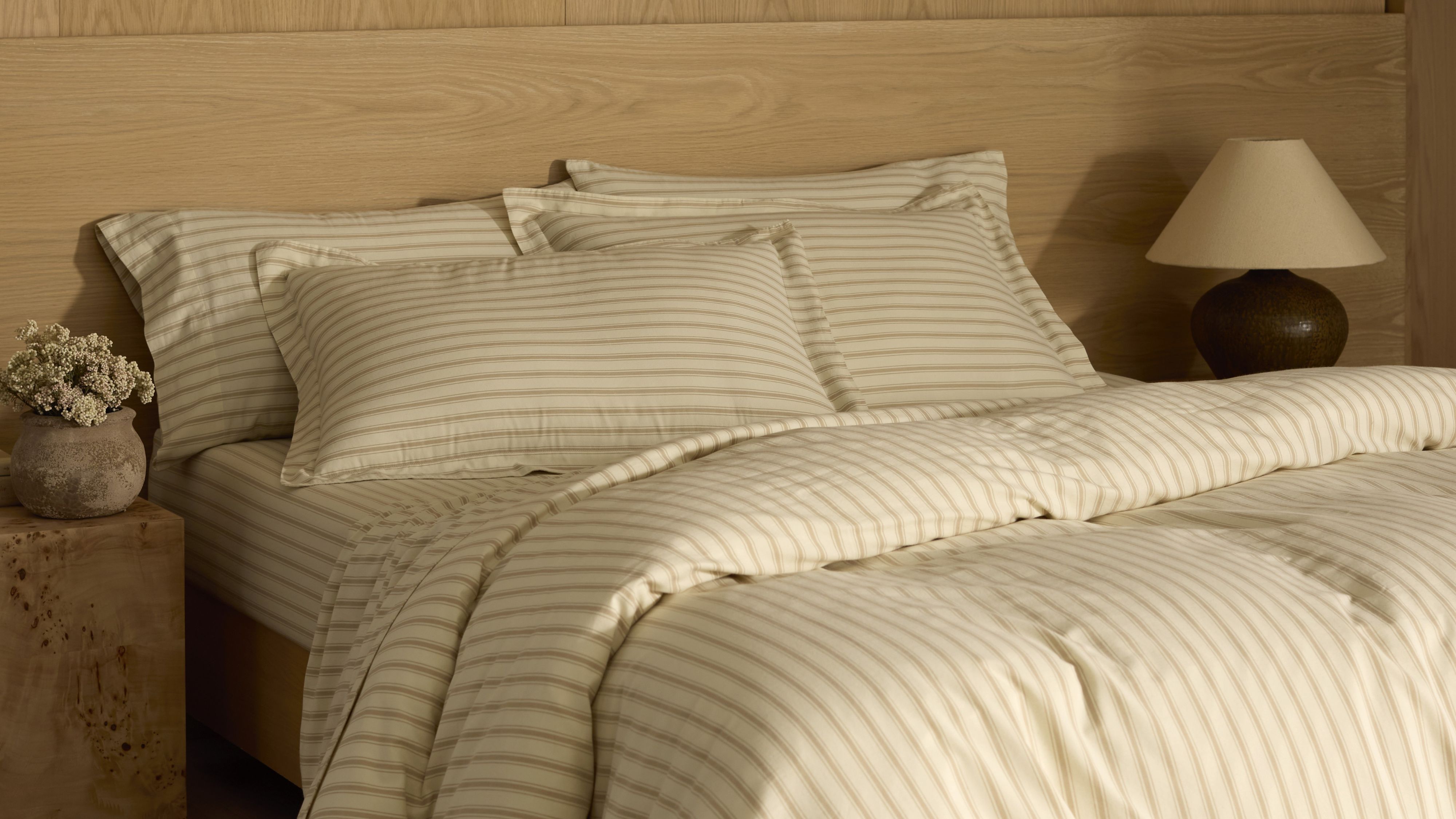Cozy Earth Soft-Wash-Bettwäsche in Kokosnuss-/Sandelholz-Streifenoptik, abgebildet in einem neutralen Schlafzimmerbereich mit einem Beistelltisch aus Wurzelholz und einem übergroßen Kopfteil mit natürlicher Maserung