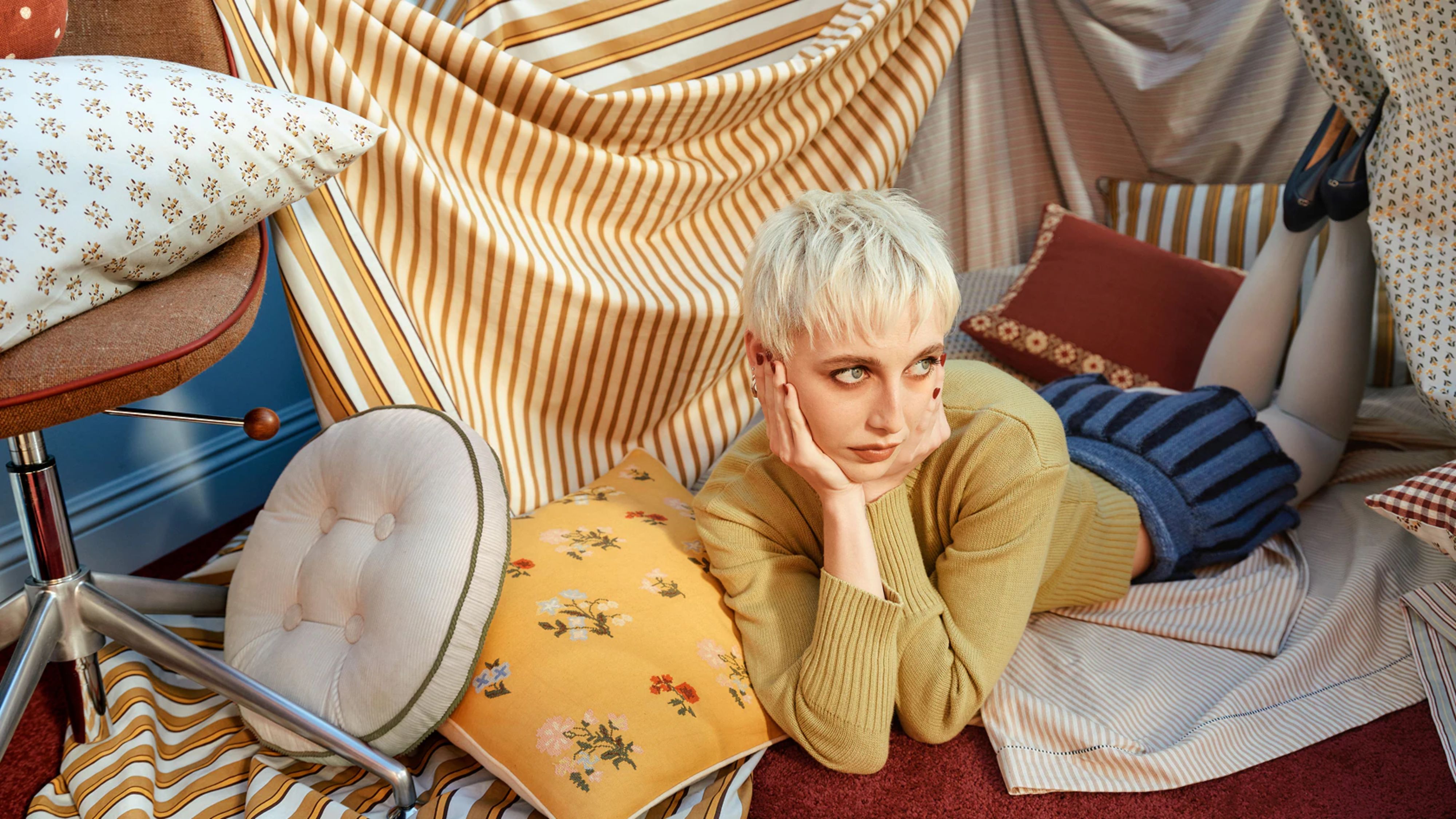 Modeikone Emma Chamberlain posiert auf dem Boden zwischen Kissen und Bettwäsche im Vintage-Stil aus ihrer neuen Wohnkollektion West Elm. 