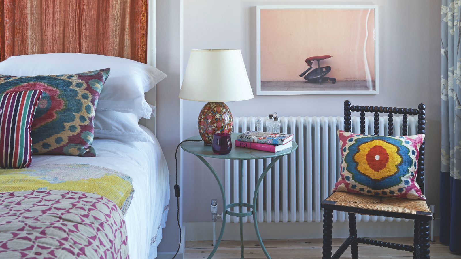 Ein farbenfrohes Schlafzimmer mit einem weißen Bett mit bunten Kissen und Decken, einem schwarzen Stuhl, einem klaren runden Tisch mit einer bunten Lampe mit weißem Schirm und einem großen Kunstwerk über dem Heizkörper.
