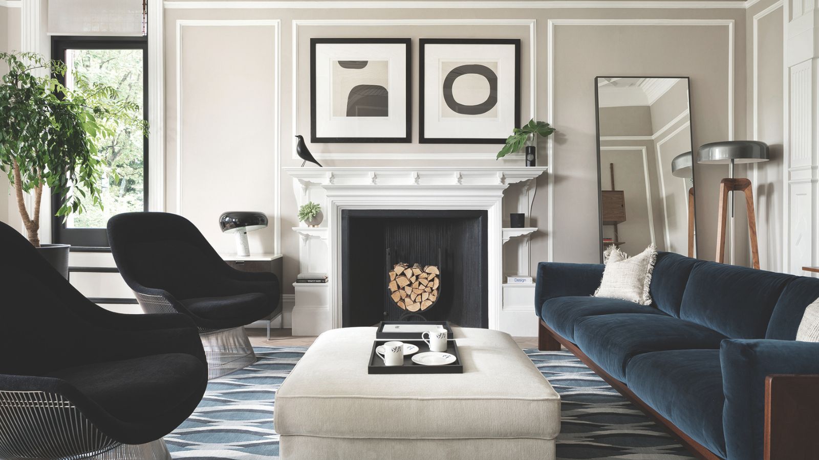 Wohnzimmer mit hellgrauen Wänden, blauem Samtsofa, zwei schwarzen Sesseln, einem hellgrauen Ottoman, einem geometrischen blauen Teppich, einem großen weißen Kamin und darüber hängender moderner Kunst.
