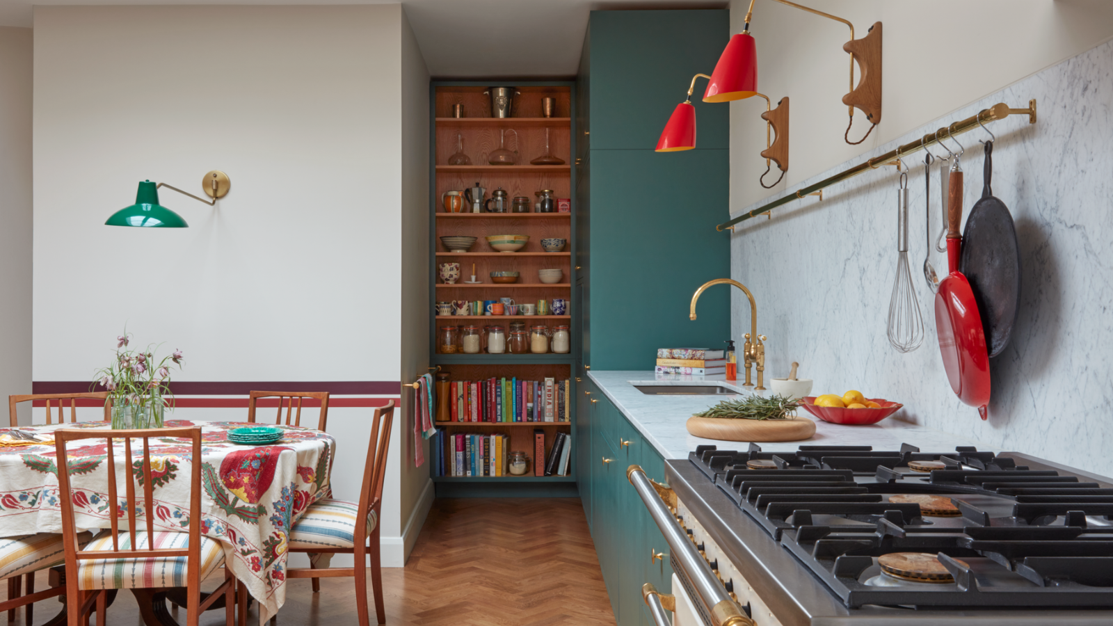 eine Küche mit einem kleinen Esstisch, einer langen Arbeitsplatte mit einem an der Wand darüber hängenden Topfregal und einem Bücherregal im Hintergrund mit Schüsseln, Gewürzen und Deko