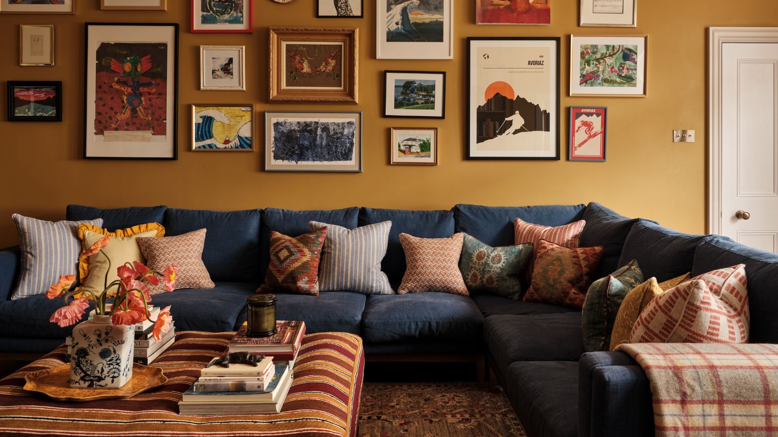 Wohnzimmer mit senffarbenen Wänden, dekoriert mit einer Sammlung von Drucken und Gemälden, einem großen dunkelblauen Sofagarnitur mit darüber verstreuten gemusterten Kissen und einem gestreiften Ottomanen in der Mitte.