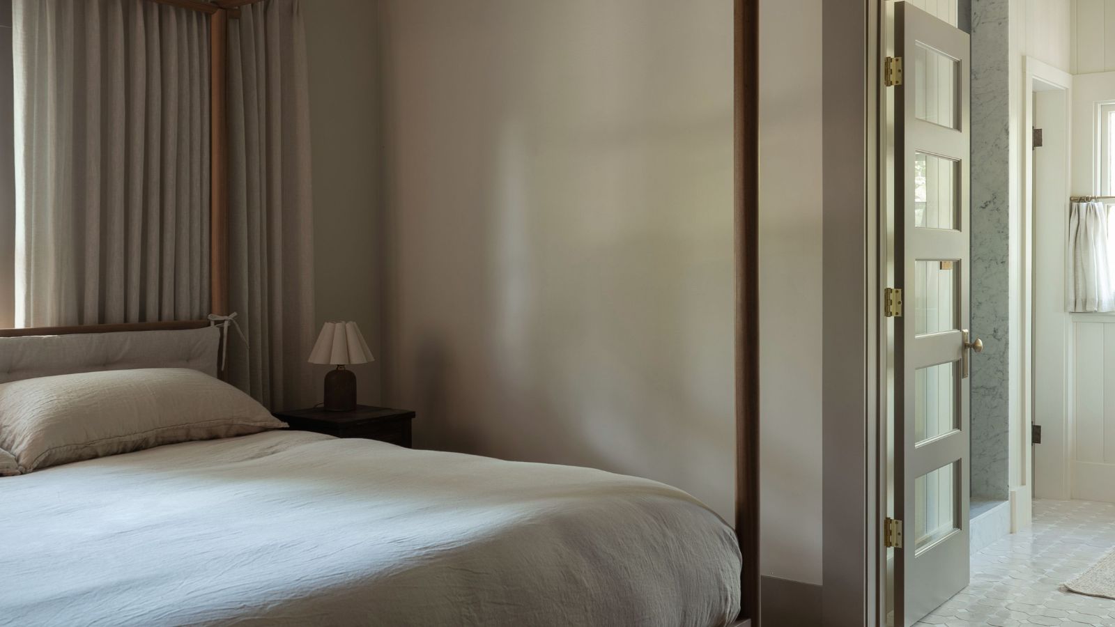 Ein modernes, beruhigendes Schlafzimmer mit taupefarbenen Wänden und dunklen Pilzbesätzen, einem minimalistischen Himmelbett mit frischer cremefarbener Bettwäsche und neutralen Vorhängen. 