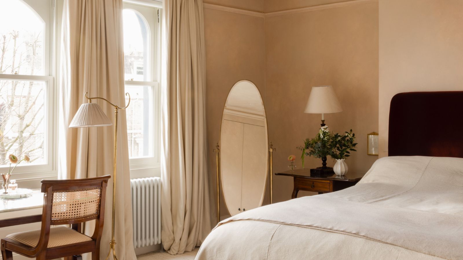 Schlafzimmer mit rosa Gipswänden, dunkelrotem Samtbett, Leinenbettwäsche, rundem Bodenspiegel, Vintage-Schreibtisch mit Rattanstuhl, Stehlampe und großen Bogenfenstern