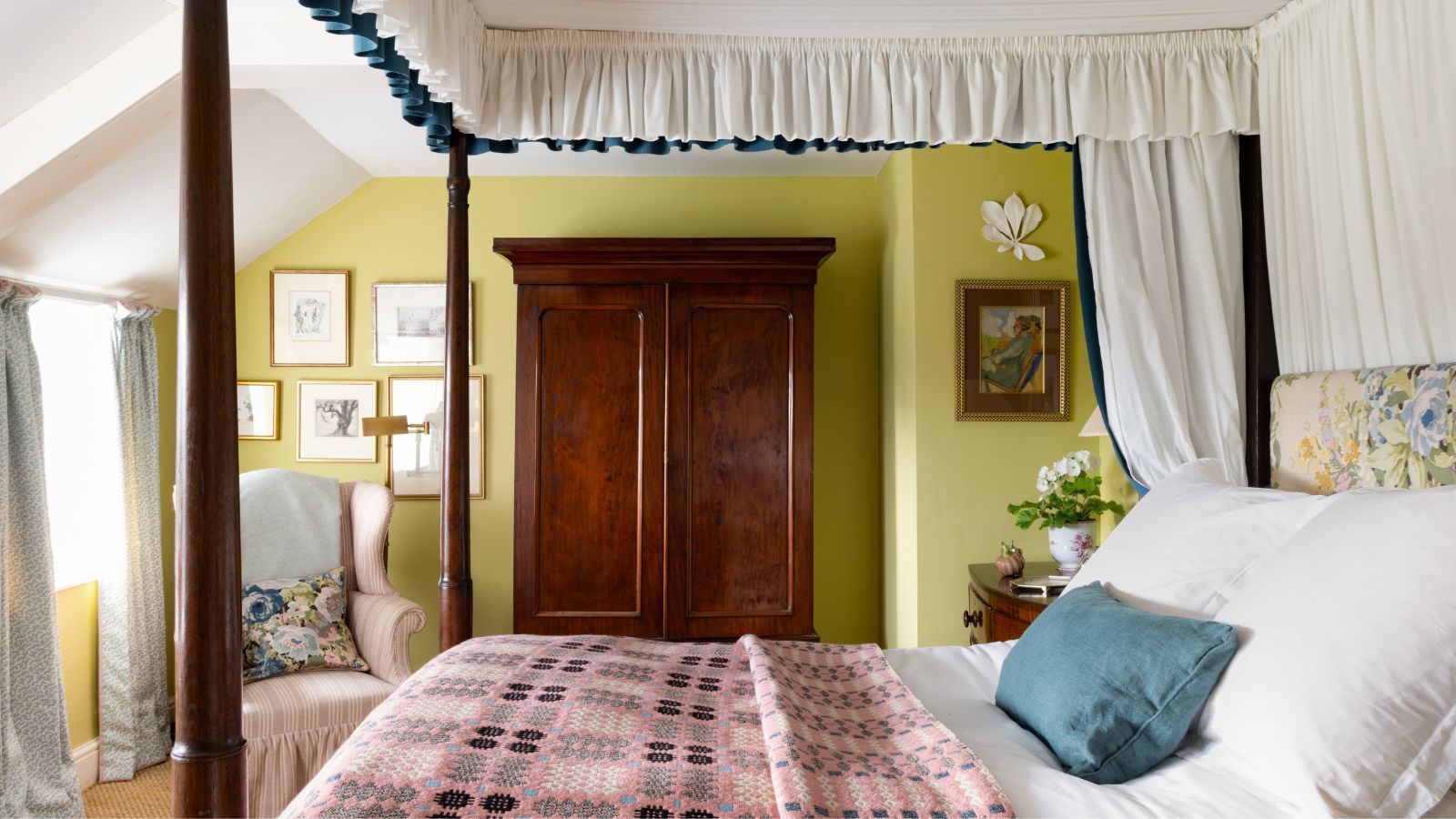 Bedroom with citron green painted walls, a mahogany four-poster bed with a canopy, a floral headboard and a pink Welsh blanket, and a large mahogany wardrobe beside a pale pink armchair.