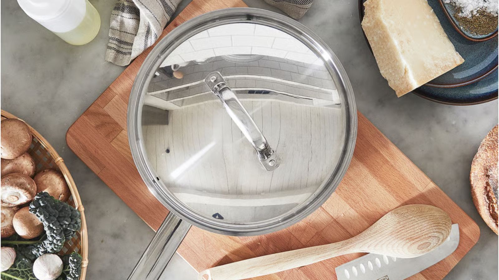 A stainless steel sauté pan with a long handle and matching lid on a wooden cutting board, viewed from above