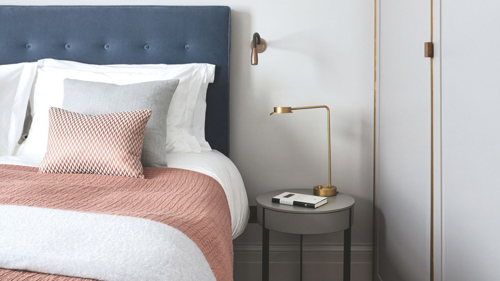 A white bedroom with a large blue bed, with white bedding, and peach blankets and cushions, white walls, and a gold lamp on a circular nightstand.