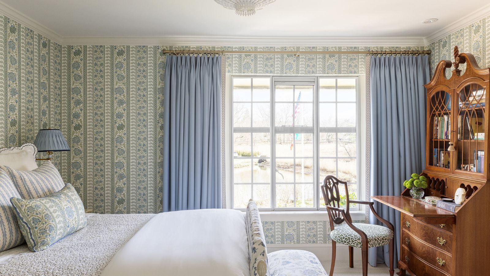 Bedroom with blue and green floral wallpaper, a large window with blue curtains, a mahogany desk filled with books and a chair underneath, and a white wooden bed with bed linen and floral cushions.