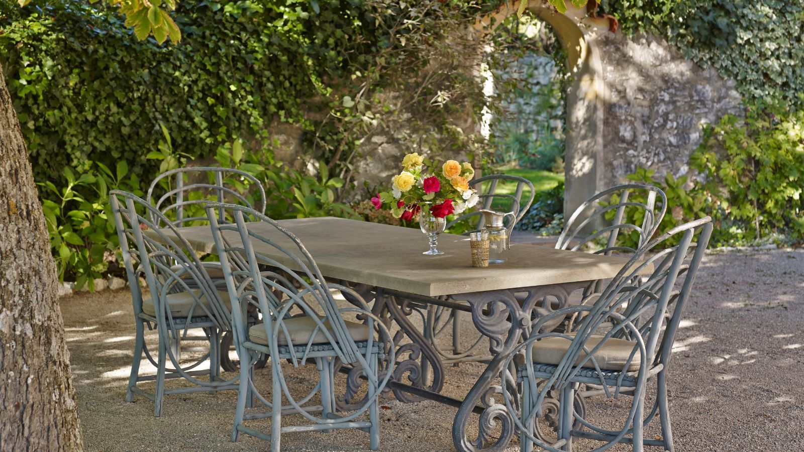 French garden with a brick arched way covered in ivy, and an antique wrought iron long dining table with vintage cane chairs underneath.