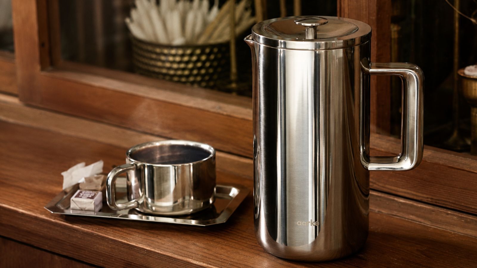 The stainless steel aarke Coffee press beside a stainless steel cup on a wooden sideboard.