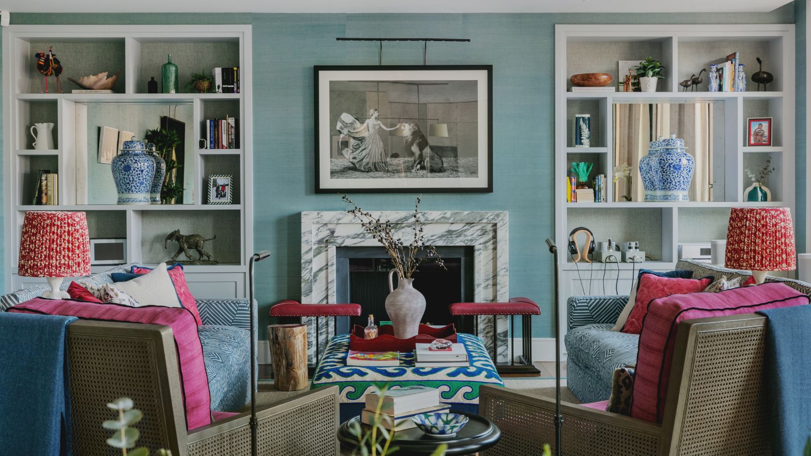 Wohnzimmer mit Wänden aus blauem Grasstoff, eingebauten Bücherregalen, Marmorkamin mit Gemälde darüber, rosa Sesseln mit roten Lampen auf beiden Seiten und zwei blauen Sofas.