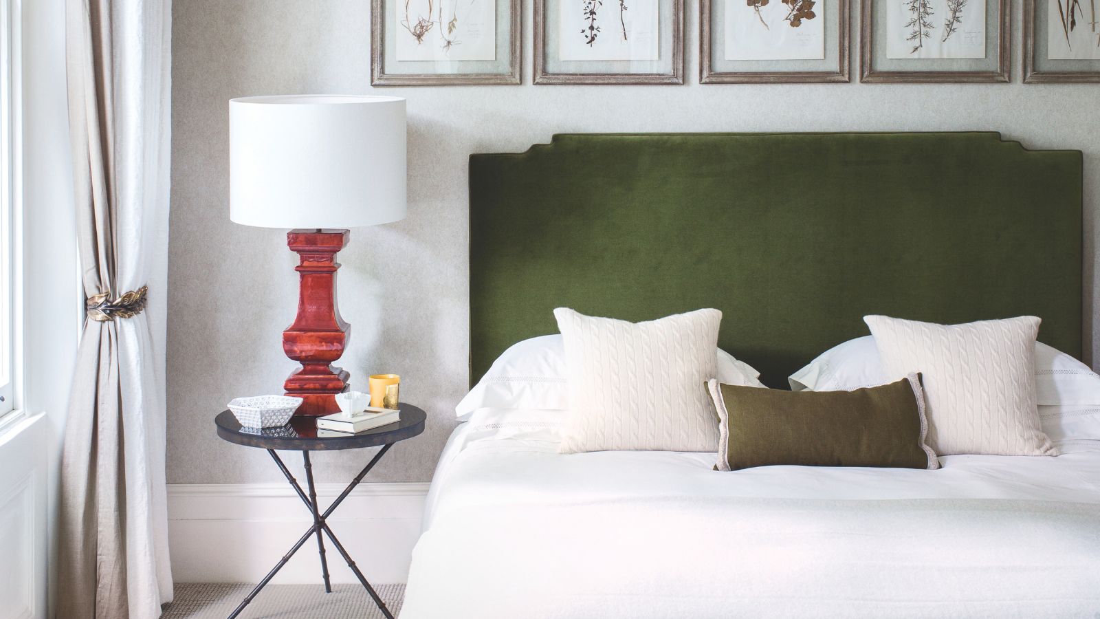 Ein neutrales Schlafzimmer mit einem Doppelbett mit grünem Samtkopfteil, weißer Bettwäsche, schwarzem Nachttisch mit roter Lampe und weißem Lampenschirm sowie fünf gerahmten botanischen Drucken über dem Bett.