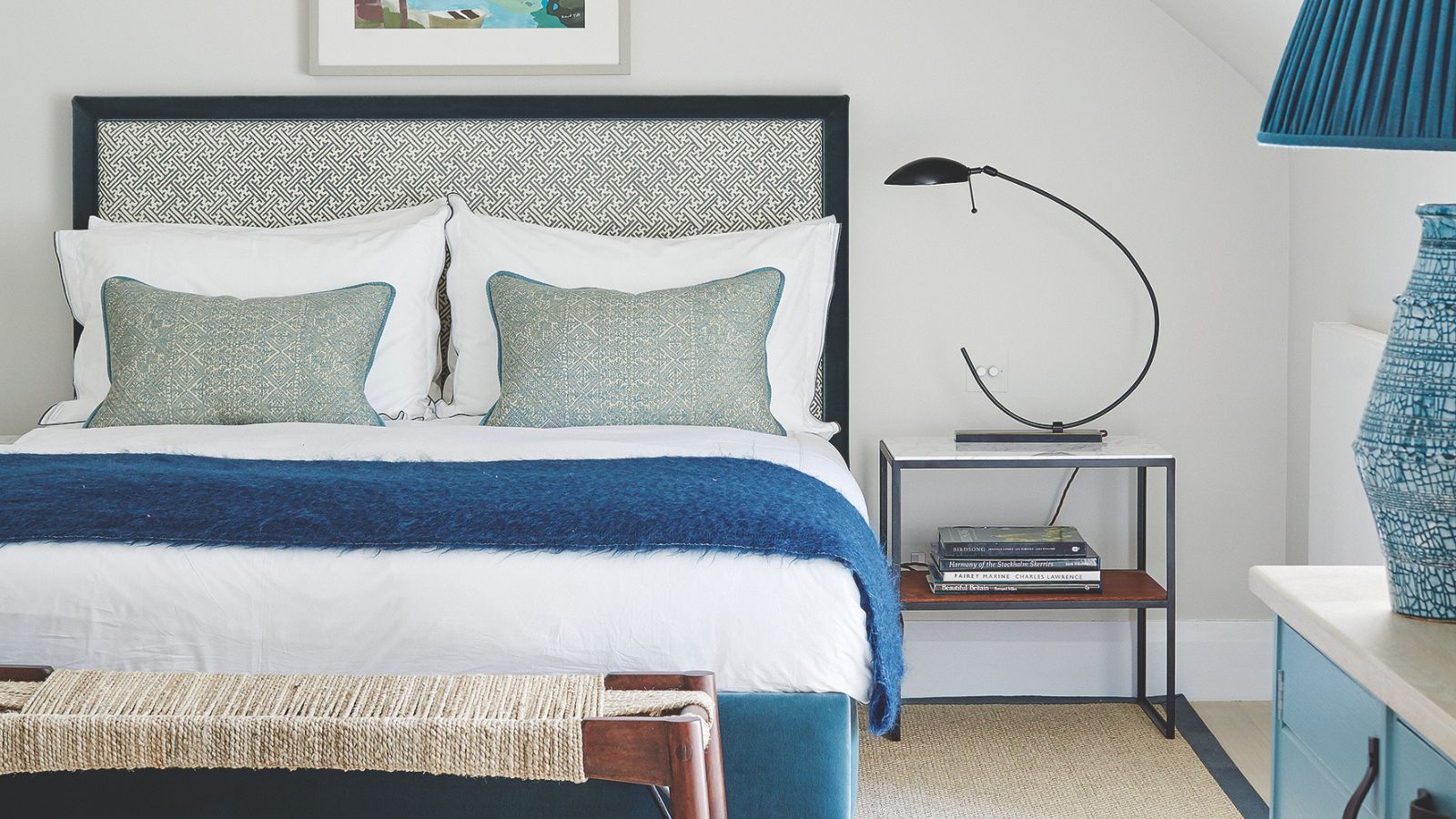 Weiß-blaues Schlafzimmer mit einem Doppelbett mit gemustertem Kopfteil, weißer Bettwäsche, blauen Decken und Kissen, weiß-blauem Nachttisch, blauer Lampe, Seil- und Holzbank und Nachttisch mit gebogener Lampe