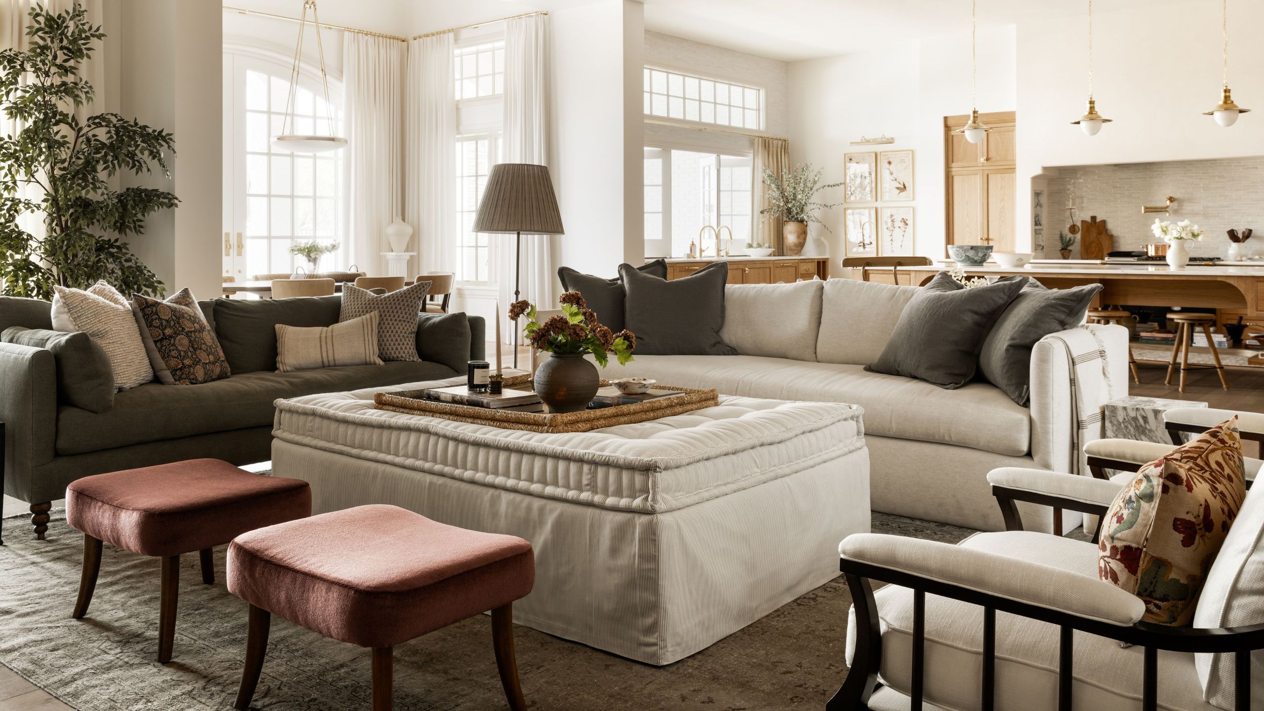 a large open plan living room kitchen area with large neutral couch, linen ottoman, rust red stools and dark green grey accents
