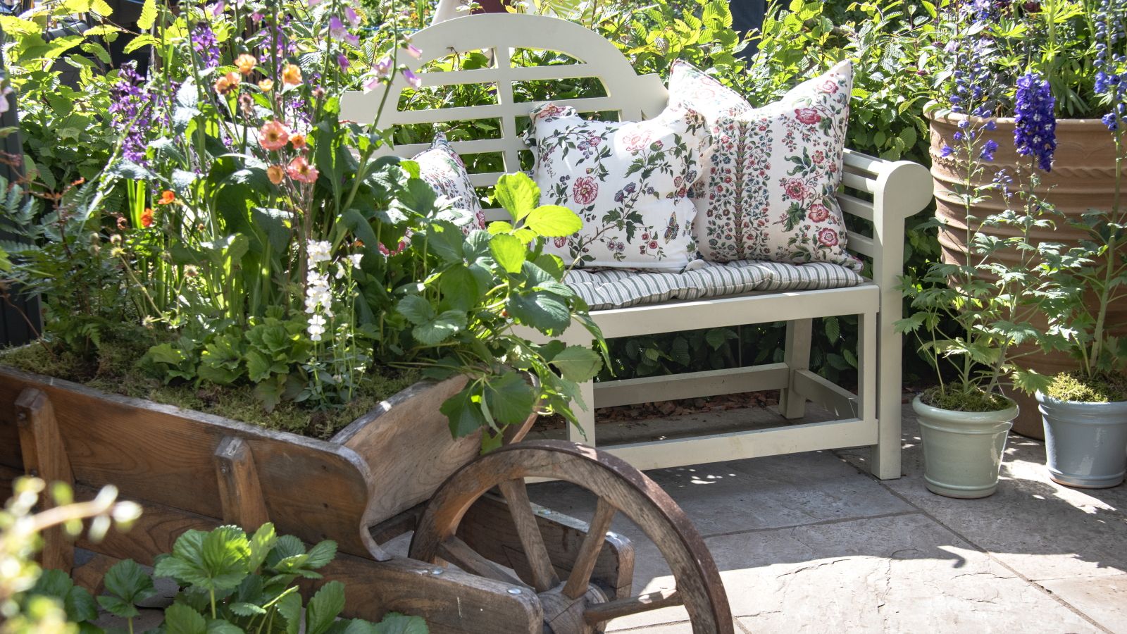An old wheelbarrow is planted up and sits on a patio next to a white bench and other containers