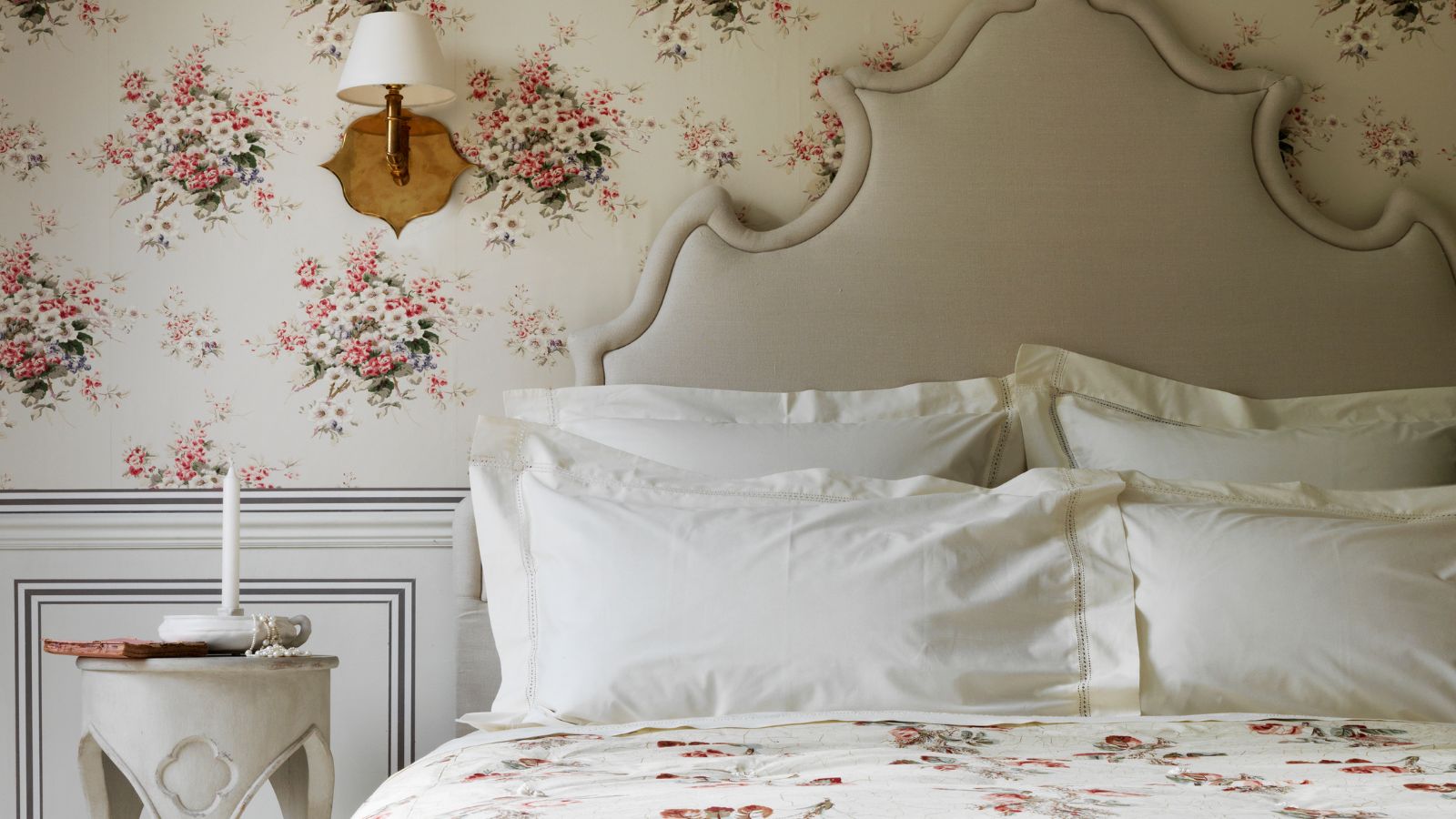 Ein neutrales Schlafzimmer mit Blumenmuster, auffälligem Kopfteil, weißen Kissenbezügen, geblümter Bettwäsche, grauer Wandverkleidung, weißer und rosafarbener Blumentapete, weißem Nachttisch und hölzerner Wandleuchte mit weißem Lampenschirm