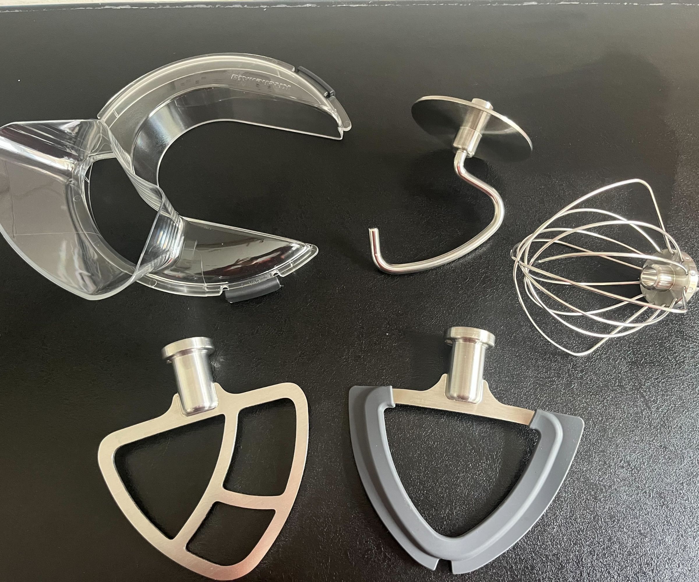 Edelstahl-Standmixer-Aufsätze auf einer schwarzen Arbeitsplatte mit einem durchsichtigen Kunststoff-Spritzschutz. Zwei Paddel, ein Schneebesen und ein Knethaken.