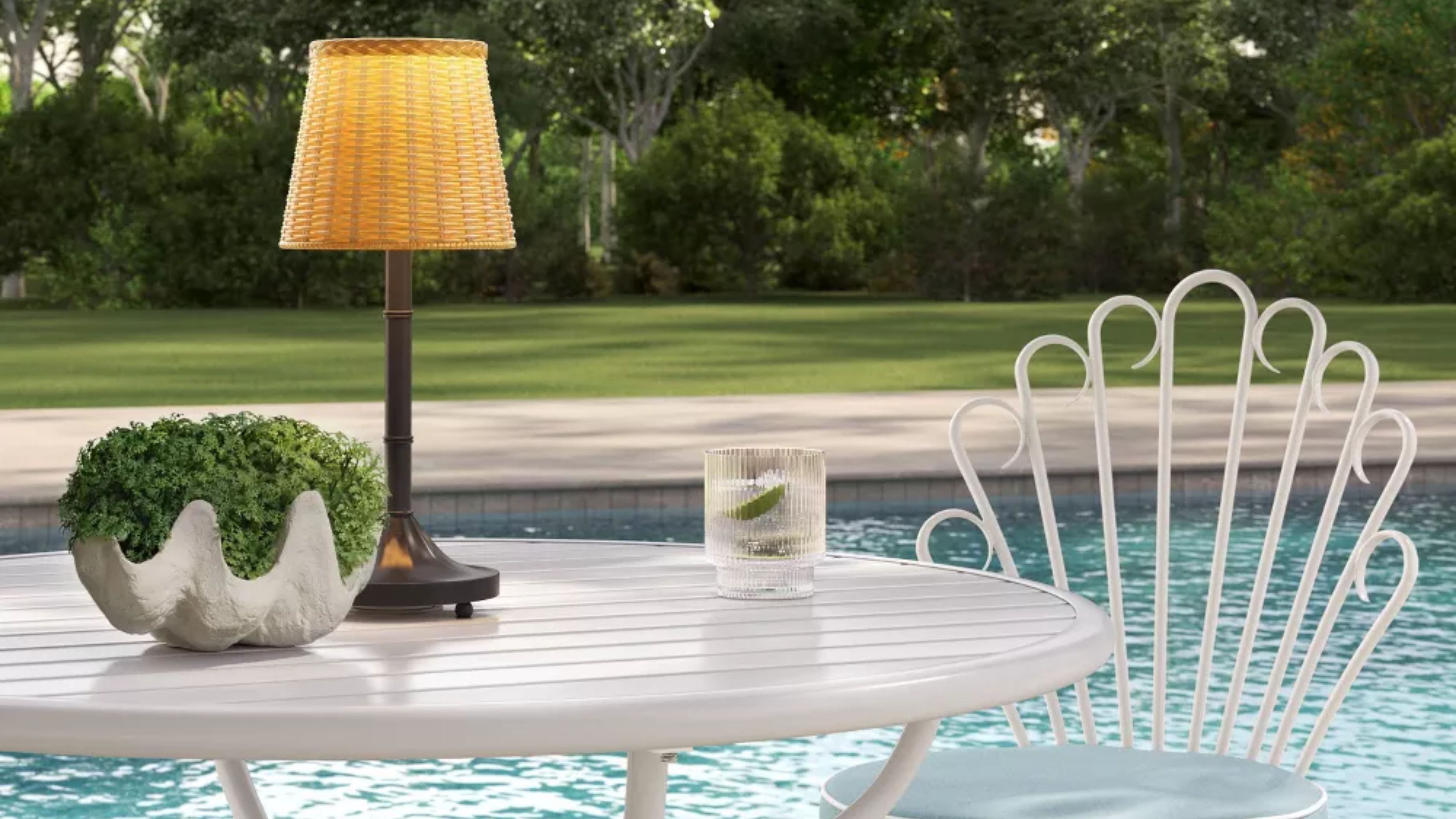 Tragbare Außenlampe aus Korbgeflecht leuchtet auf einer weißen Terrasse mit Blick auf einen Pool und eine Rasenfläche