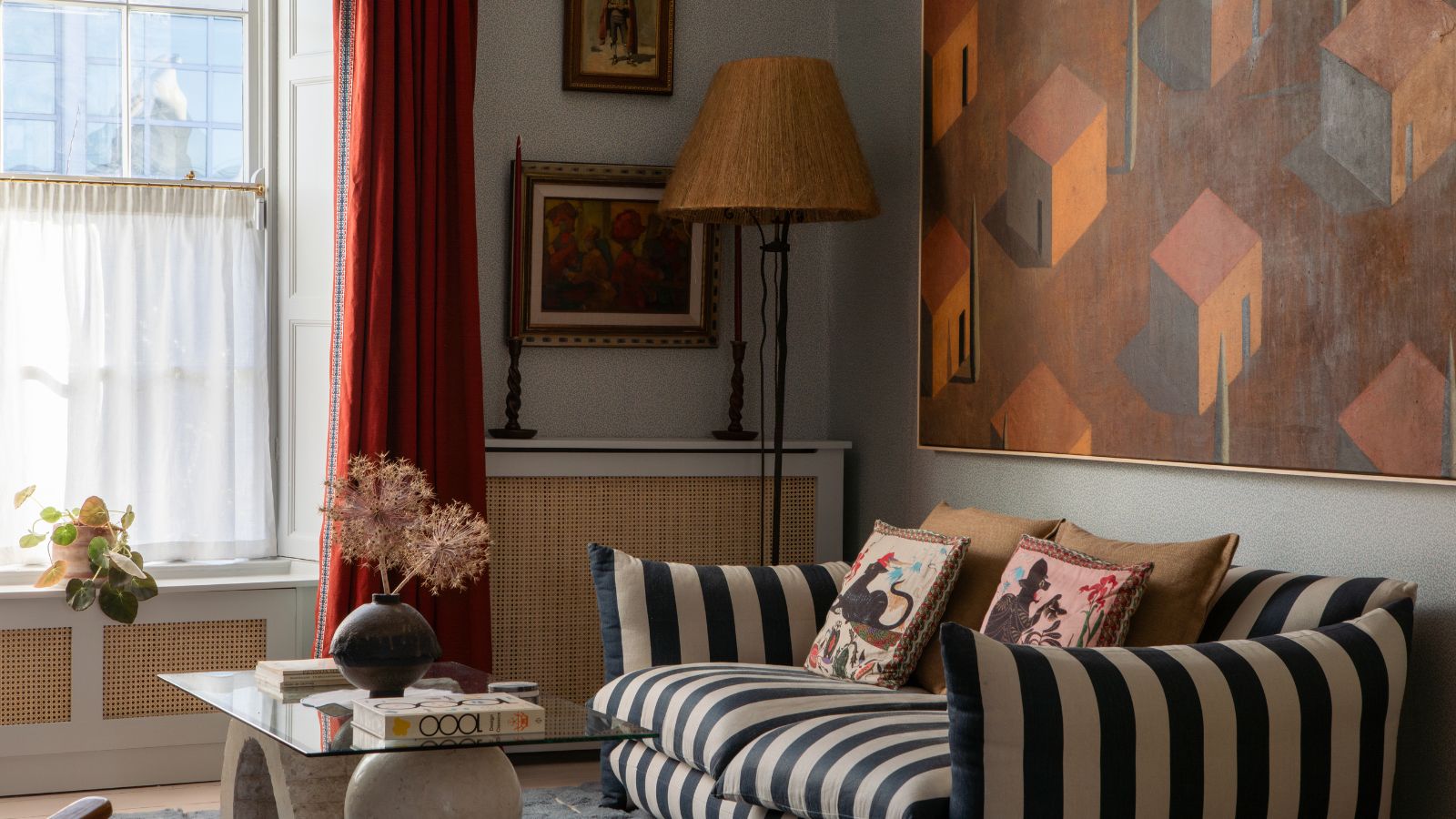 Wohnzimmer mit hellblauen Wänden, gestreiftem Sofa, roten Vorhängen, einem Couchtisch aus Glas und Stein, einem großen Gemälde über dem Sofa in Orange und Rot mit Häusern und einer Stehlampe aus Rattan in der Ecke