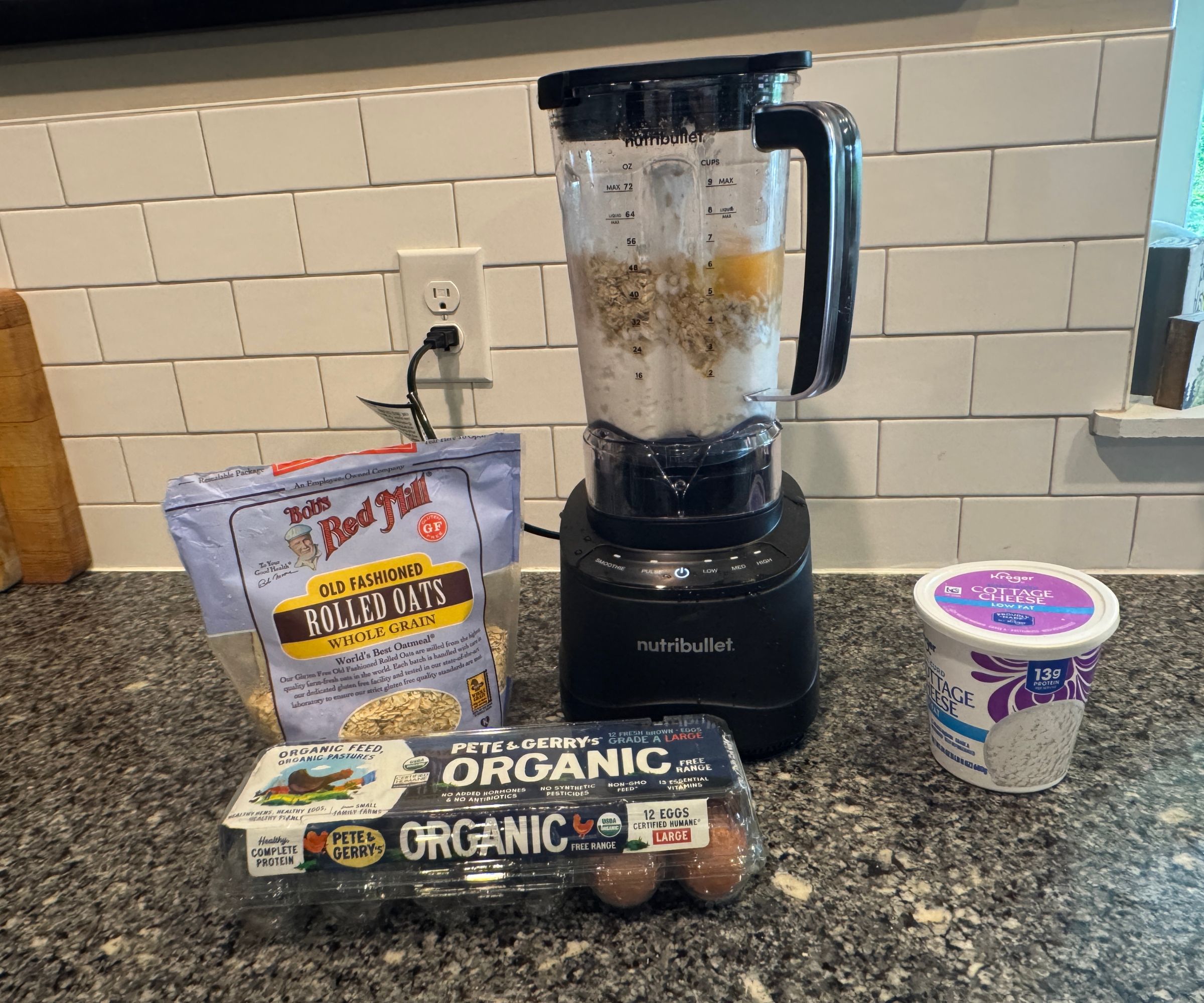 Ein schwarzer Mixer, umgeben von Eiern, Haferflocken und Hüttenkäse.