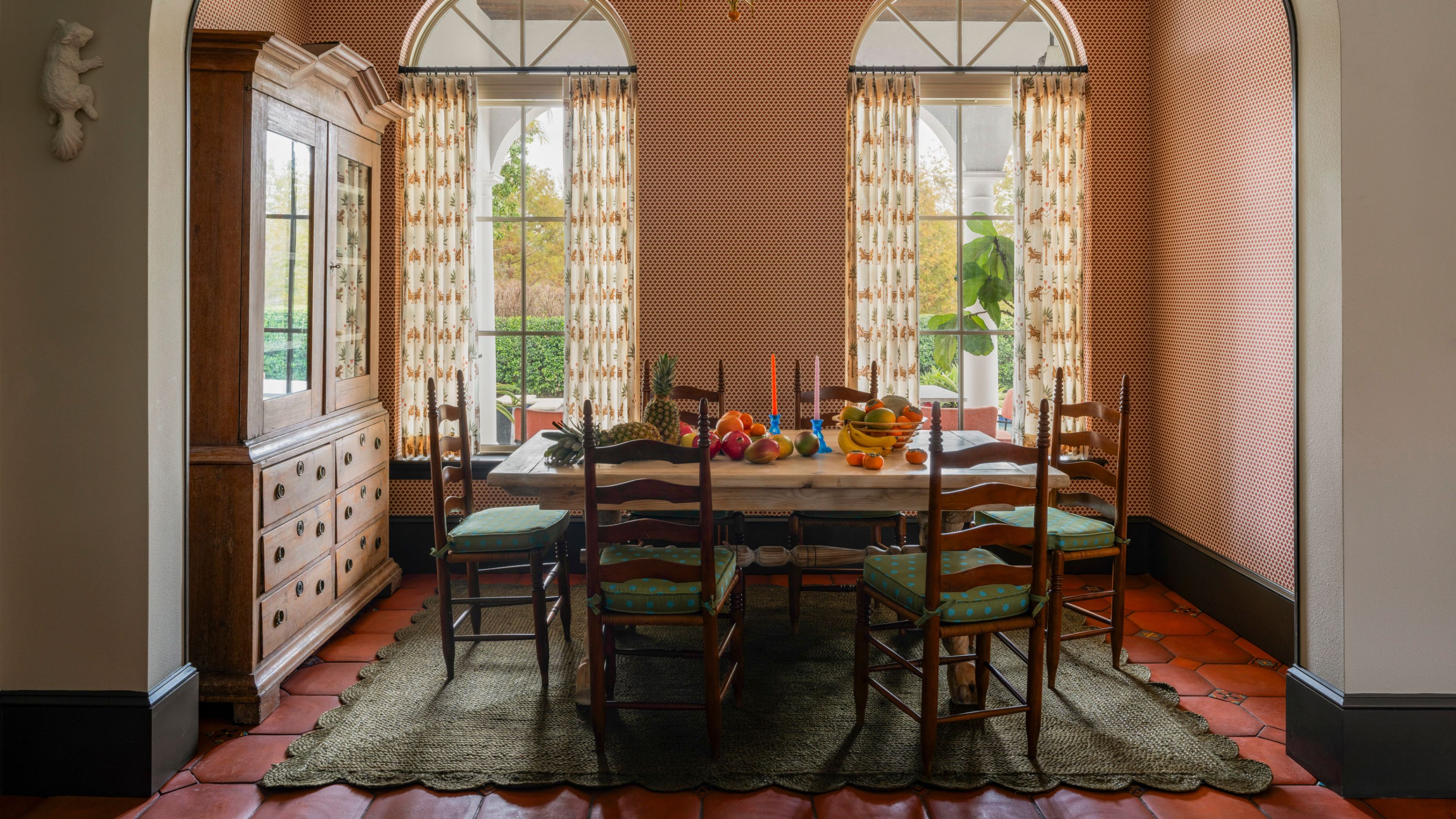 Ein sonniges Esszimmer in einem Haus in Miami mit großen gewölbten Terrassentüren, tapezierten Wänden, Terrakottaböden, einem grünen Wellenteppich und antiken Möbeln