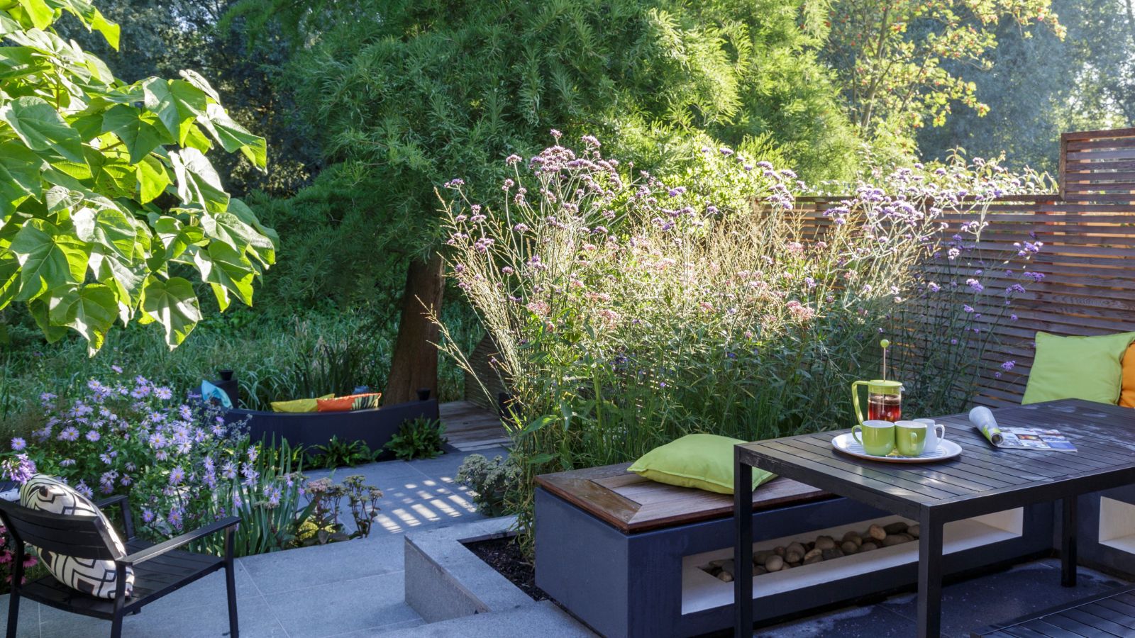 Terrassensitzbereich im sonnigen Garten mit Tisch, Sitzbänken, Kaffeemaschine, Kaffeetassen und bunten Kissen. Hinter der Terrasse stehen ein schwarzer Stuhl mit einem schwarz-weißen Kissen, Blumen und Bäume.