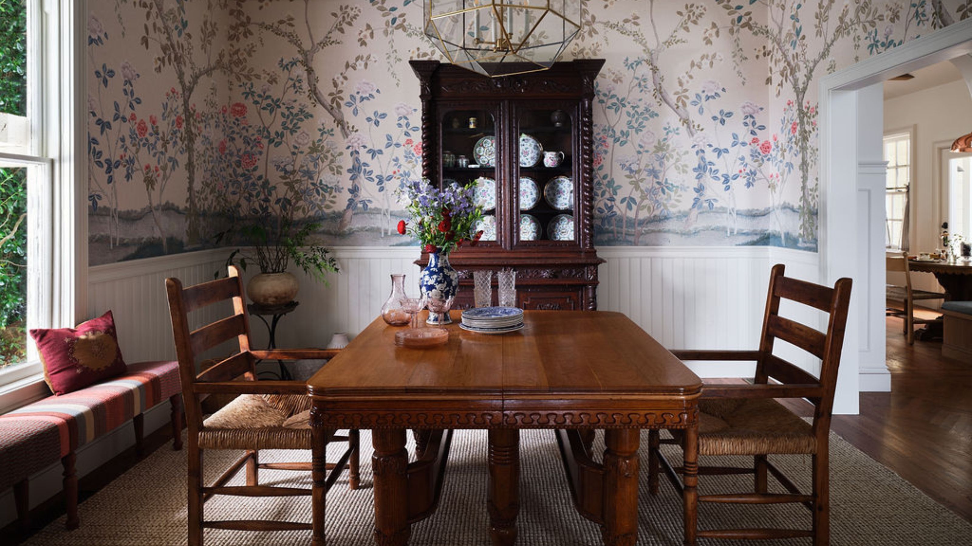 Ein Esszimmer mit rosafarbener Blumenwandtapete, halbhoher Täfelung, antiken Möbeln aus dunklem Holz und einer Fensterbank
