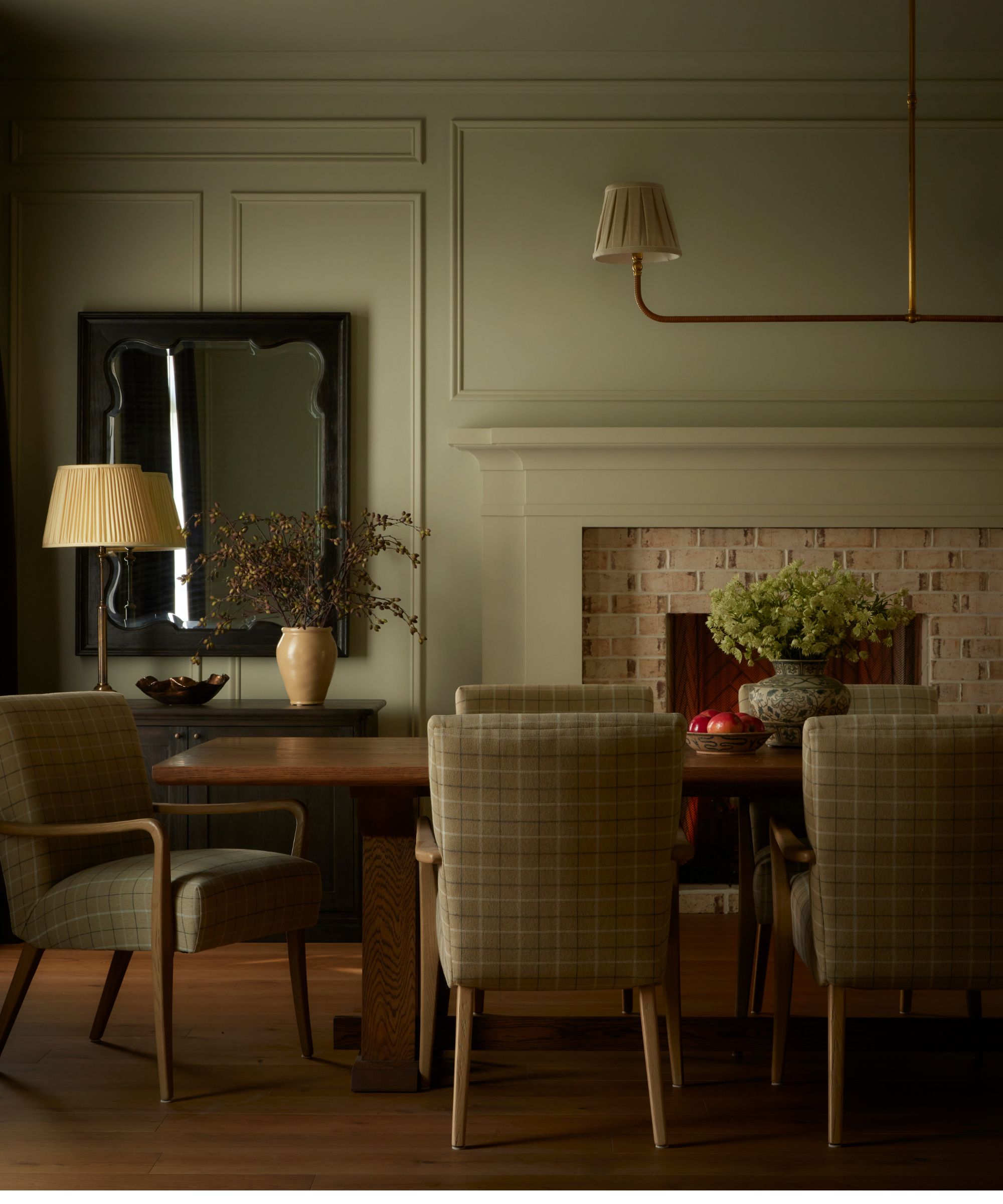 Gemütliches Esszimmer mit dunklem rechteckigem Esstisch aus Holz, gepolsterten Esszimmerstühlen und einer Blumenvase auf dem Tisch. Gemauerter Kamin und Sideboard mit Lampe und Vase im Hintergrund.