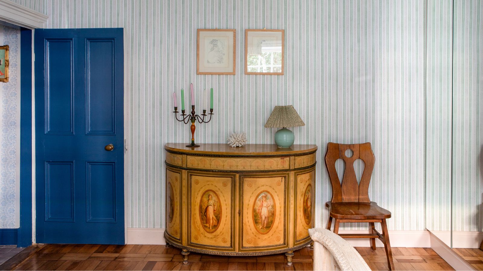 Schlafzimmer mit einer blau gestrichenen Tür, gestreifter blauer Tapete, einem Vintage-Konsolentisch mit bemaltem Design, einem eisernen Kandelaber darauf mit bunten Kerzen und einem Vintage-Stuhl daneben