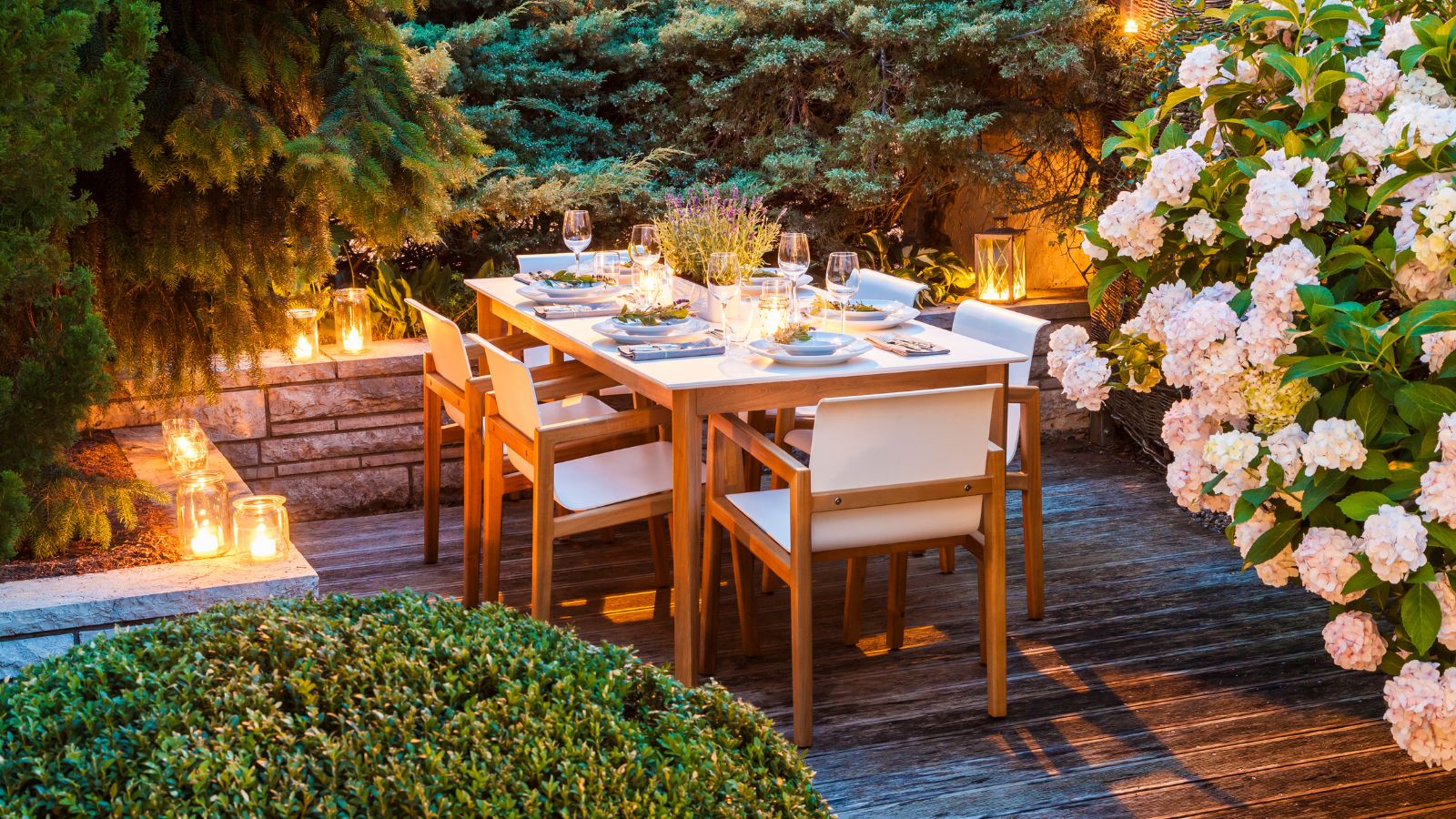 Terrasse mit hölzernem Esstisch, der für das Abendessen gedeckt ist, umgeben von viel Grün und beleuchtet von leuchtenden Kerzen