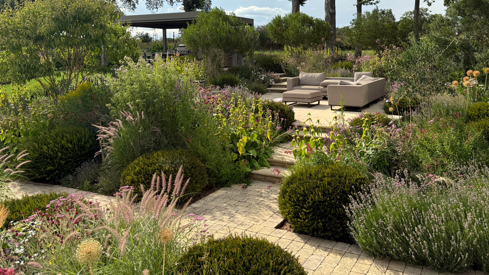Gartengestaltung auf zwei Ebenen mit Pflanzbeeten, erhöhtem Terrassenbereich mit Gartenmöbeln und Pergola, alles durch gemauerte Wege verbunden