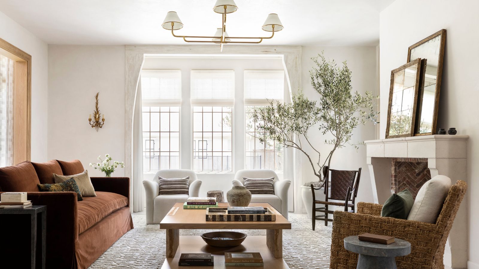Ein helles und luftiges Wohnzimmer mit weißen Wänden, großen Fenstern, einem rostfarbenen Sofa und einem großen Couchtisch aus Holz mit Büchern und Deko. Ein strukturierter Teppich und zwei cremefarbene Sessel. 