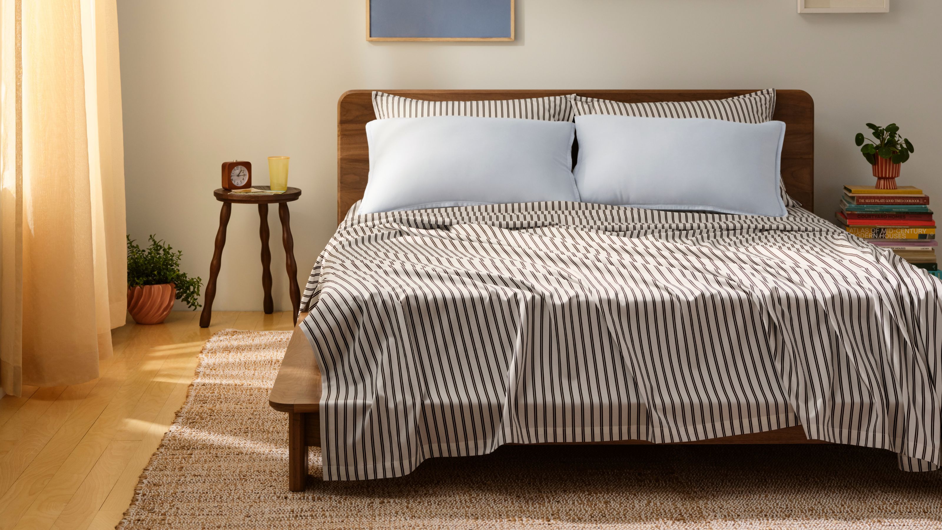 Gestreifte braun-weiße Brooklinen-Tencel-Laken mit blauen Kissen auf einem hölzernen Doppelbettgestell in einem weißen Schlafzimmer mit hellem Holzboden, Juteteppich, rundem Nachttisch, Büchern und pfirsichfarbenen Vorhängen.