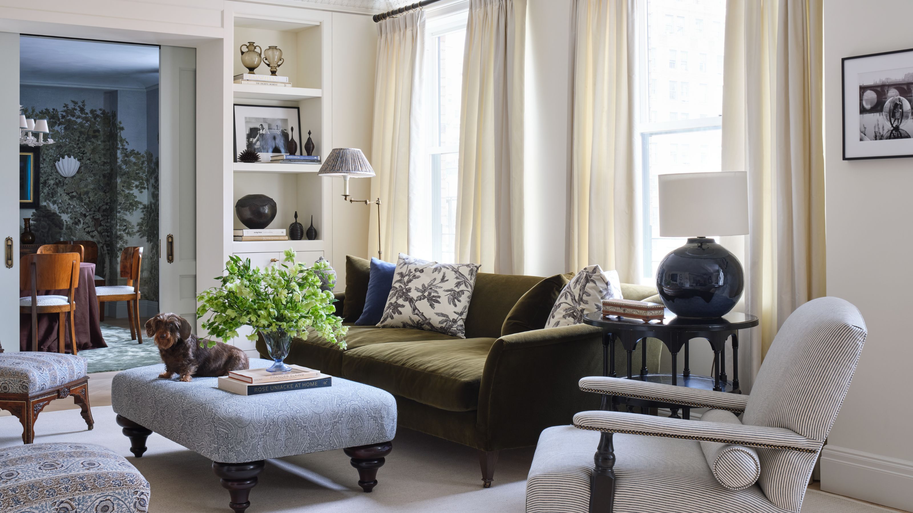 Ein großes, formelles Wohnzimmer in New York mit eleganten Zierleisten, neutralen Wänden, Bücherregalen am Eingang, einem olivgrünen Samtsofa, kleinen Akzentstühlen und einem Ottoman