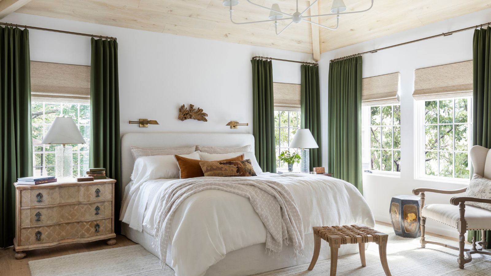 Ein modernes, helles und luftiges Schlafzimmer mit heller Holzdecke, weißen Wänden, einem großen beigen Teppich, olivgrünen Vorhängen und einem großen Bett mit mehrlagiger neutraler Bettwäsche. 