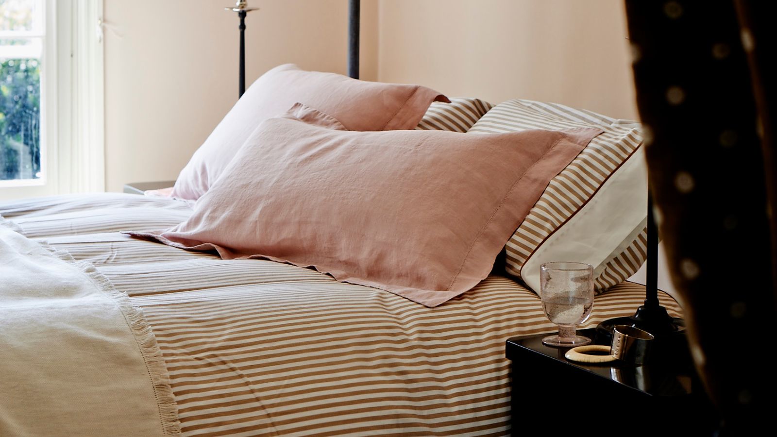 Ein pfirsichfarbenes Schlafzimmer mit gestreifter rosa Bettwäsche, rosa Kissen und einem dunkelbraun-weiß gepunkteten Vorhang im Vordergrund.