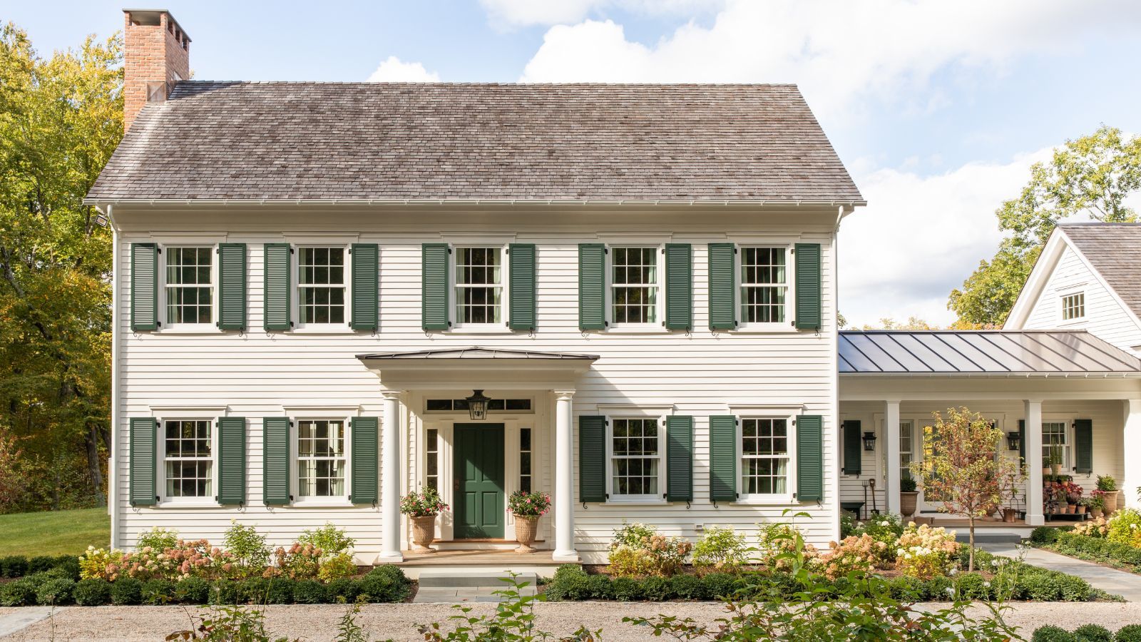 A large traditional house exterior with warm white siding, a dark green front door and dark green shutters, with a porch and flower beds at the front of the home.