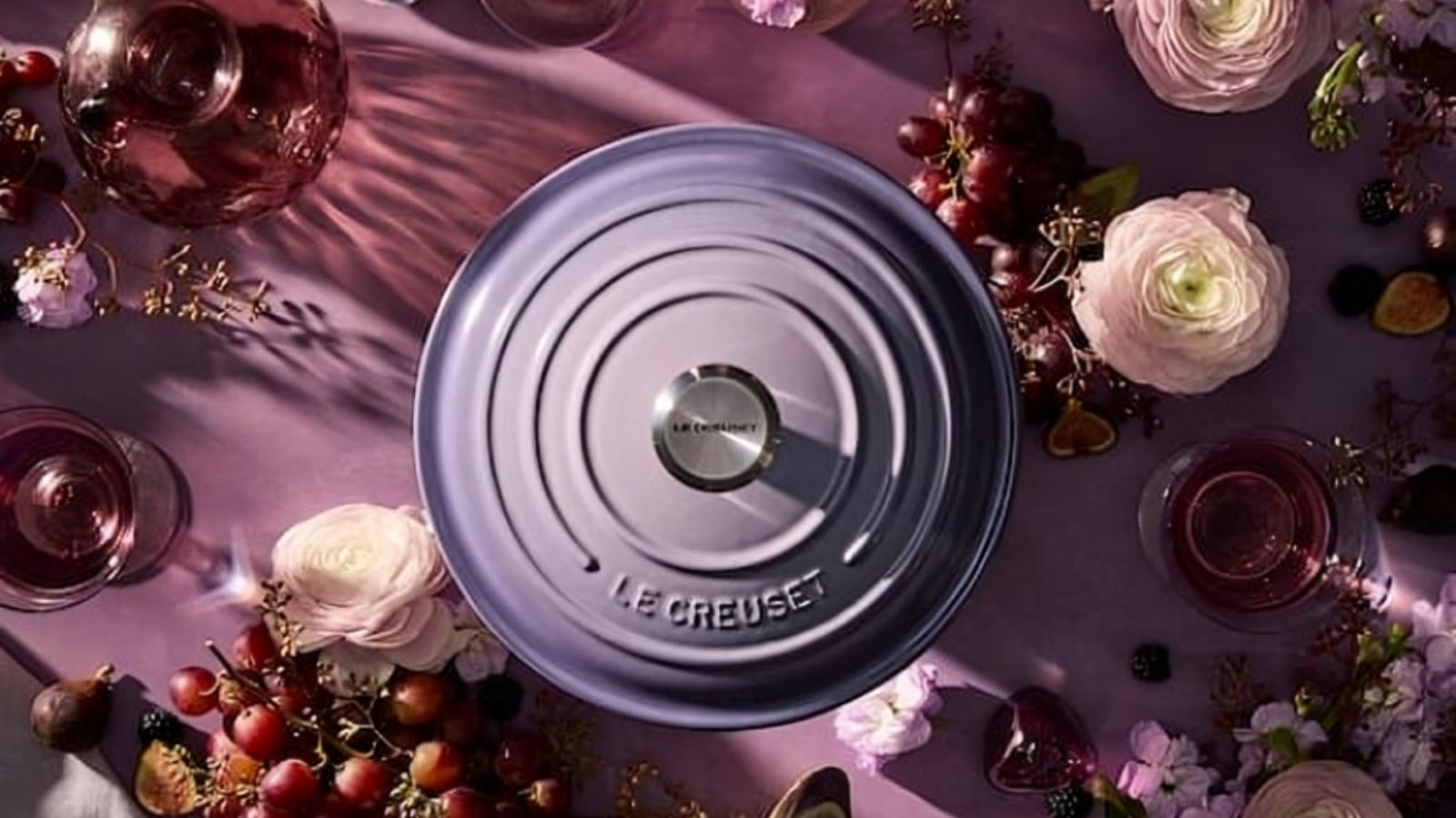 Ein violetter Deckel eines holländischen Ofens von Le Creuset, von oben betrachtet, auf einer violetten Arbeitsplatte, umgeben von violetten und hellrosa Blumen
